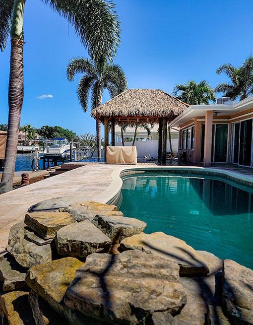 Swimming pool beside a waterway, thatched-roof gazebo, palm trees under a blue sky.