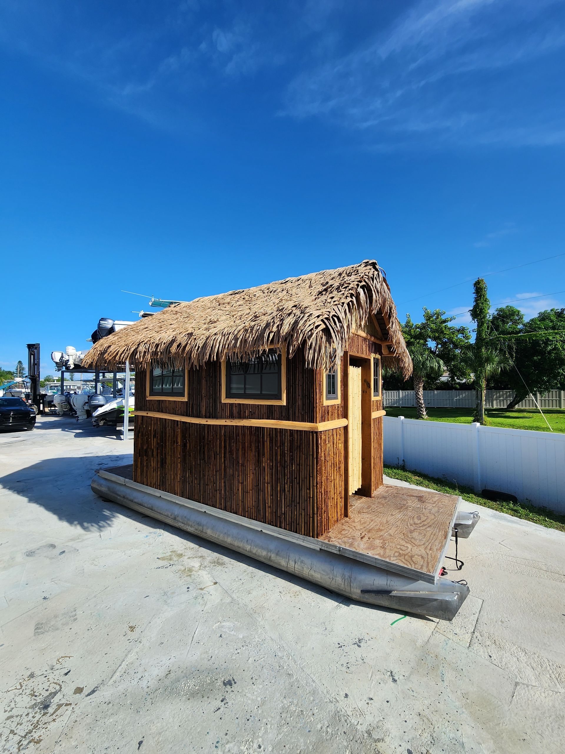 Wooden deck attached to a house, with stairs leading down to an above-ground pool. Wooden hut with thatched roof, on a pontoon, under a blue sky.