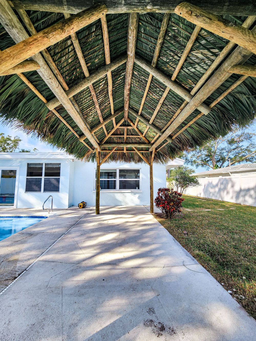 Underneath a thatched roof, a concrete walkway leads to a light blue house beside a pool.