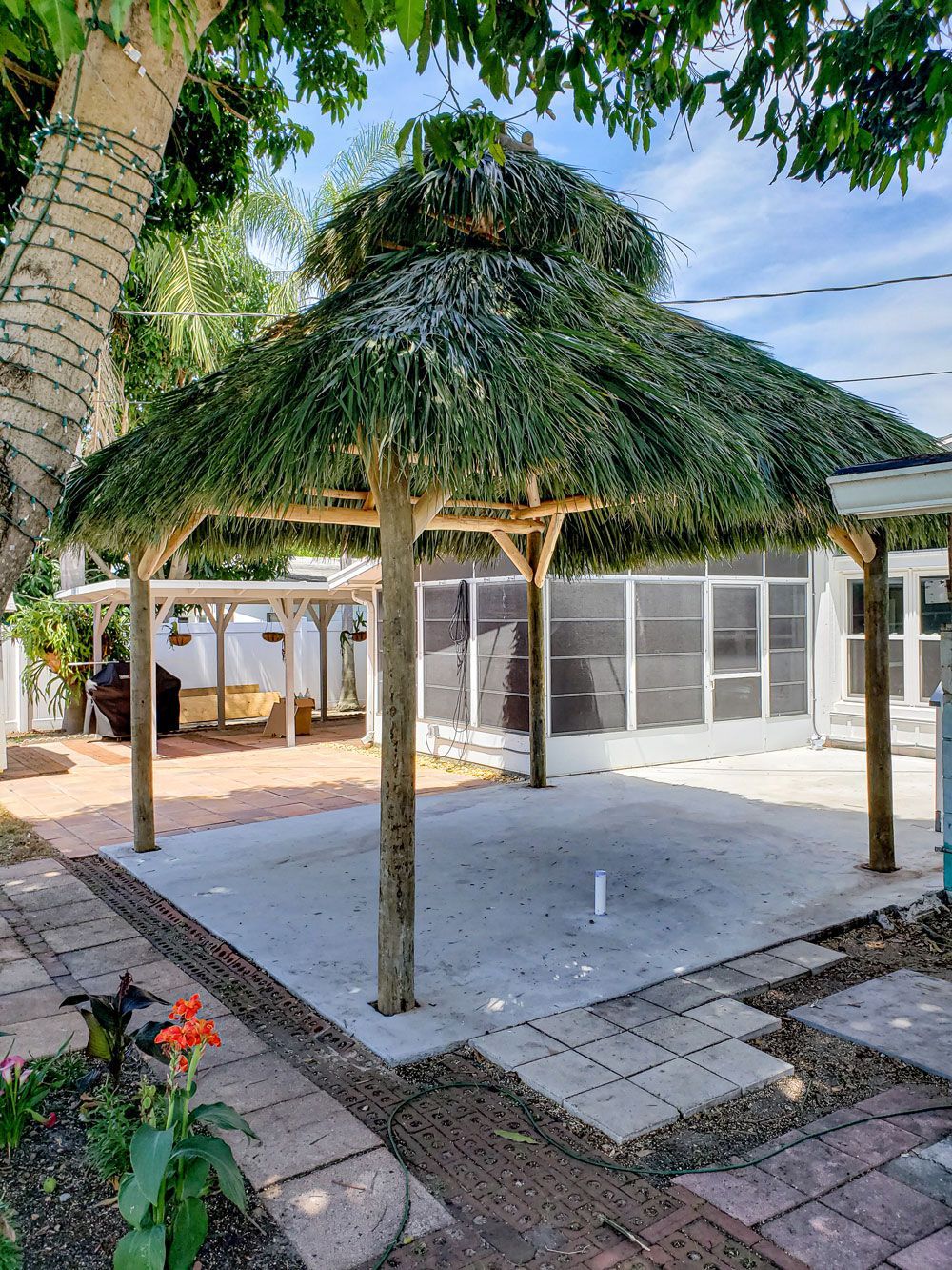 A thatched roof gazebo on a concrete patio, surrounded by brick and greenery.