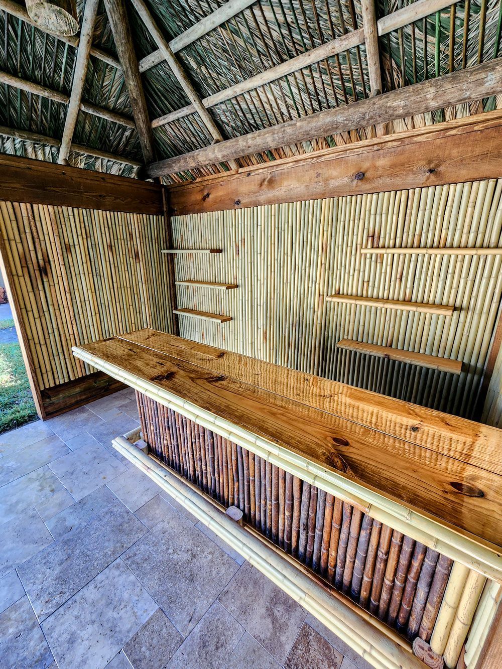 Bamboo-decorated outdoor bar with wooden counter and shelves, thatched roof.