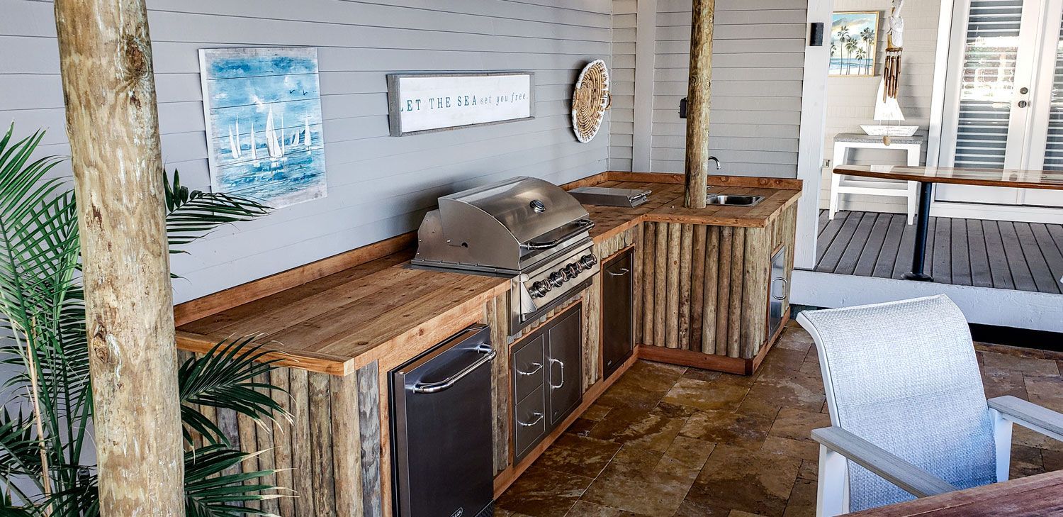Outdoor kitchen with wooden countertops, grill, refrigerator, and log siding. Beach-themed wall decor.