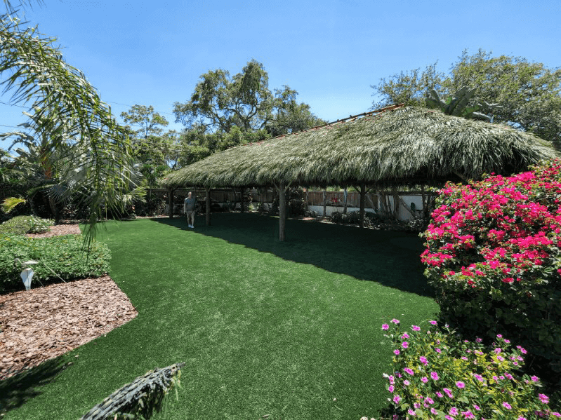 A large thatched-roof structure with a green lawn and flowering bushes under a blue sky.