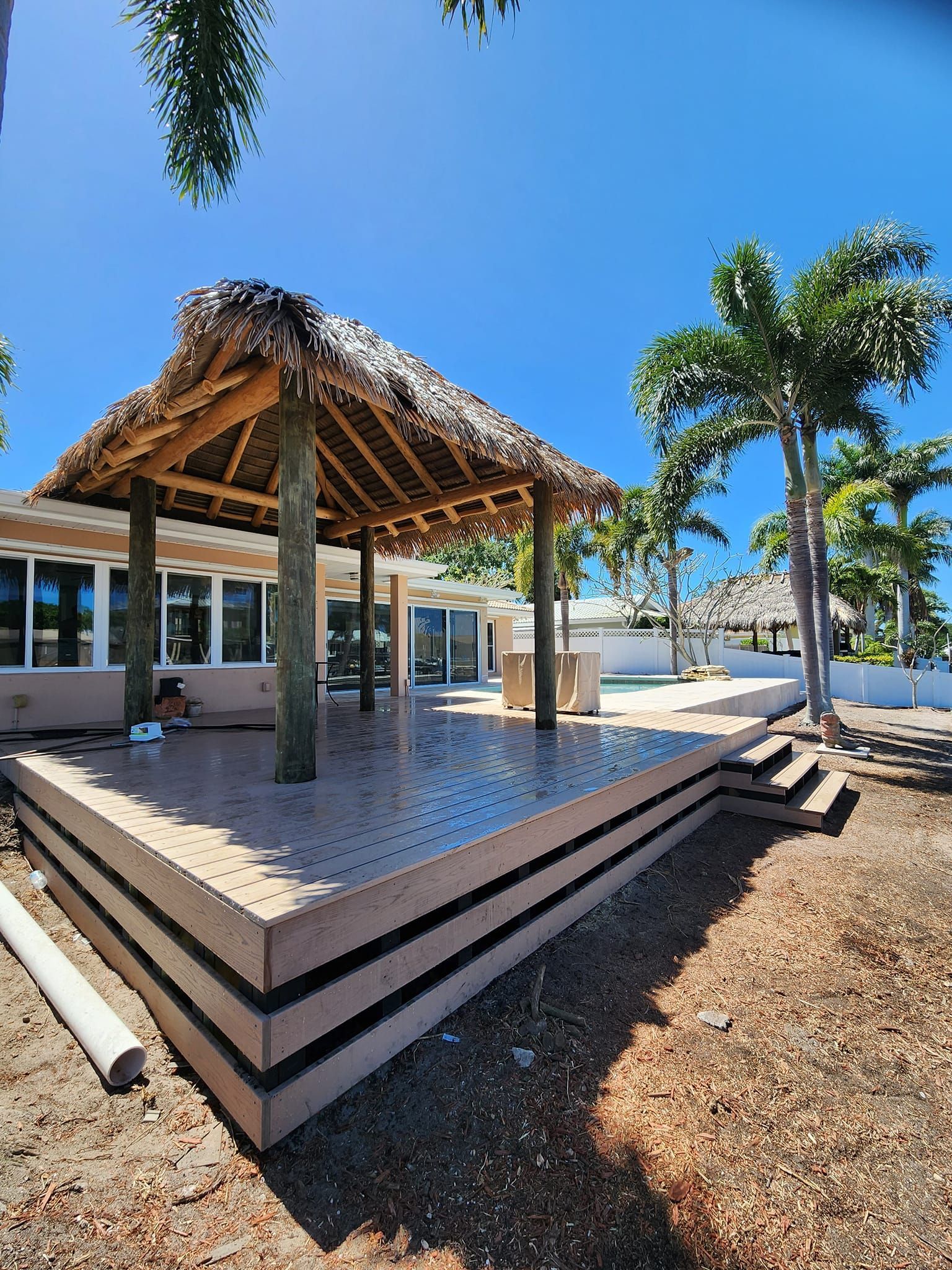 Wooden tiki hut on a raised concrete patio, next to a building with windows, under a blue sky. Wooden tiki hut on a raised concrete patio, next to a building with windows, under a blue sky.