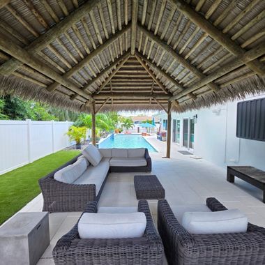 Outdoor seating area under a thatched roof, with a pool visible in the background.