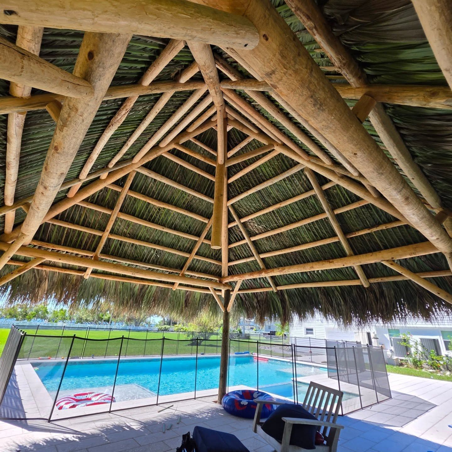 Tiki-style roof over a pool with a safety fence. Wooden beams, green thatched roof, blue water, and outdoor seating. Tiki-style roof over a pool with a safety fence. Wooden beams, green thatched roof, blue water, and outdoor seating.