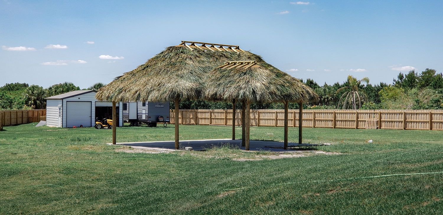 Tiki-style hut with a thatched roof, set in a grassy yard, with a fence and a shed in the background.