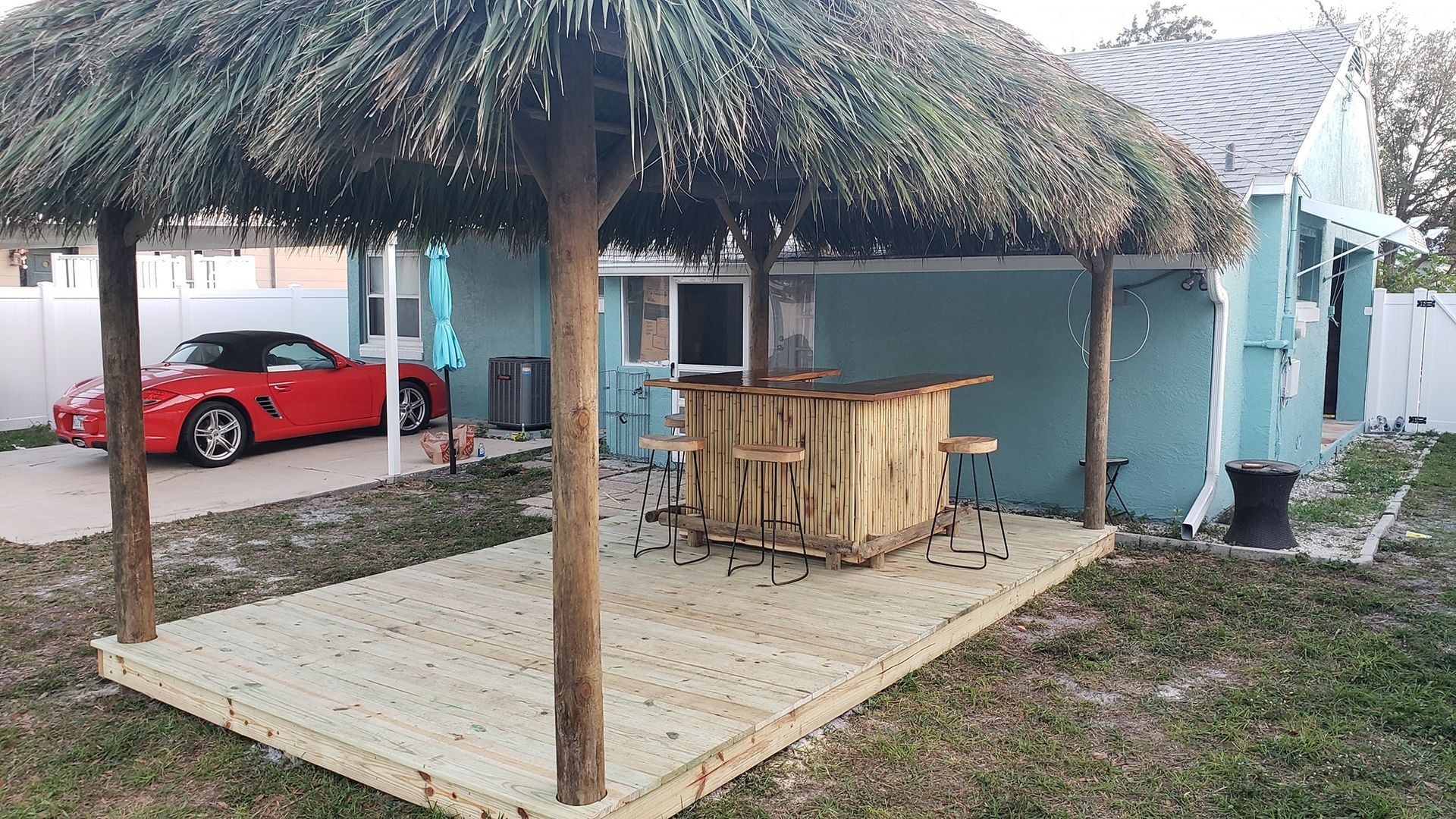 Tiki bar with thatched roof and wooden deck in backyard; red car in background. Tiki bar with thatched roof and wooden deck in backyard; red car in background.