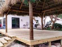 Wooden deck with thatched roof gazebo attached to a white house.