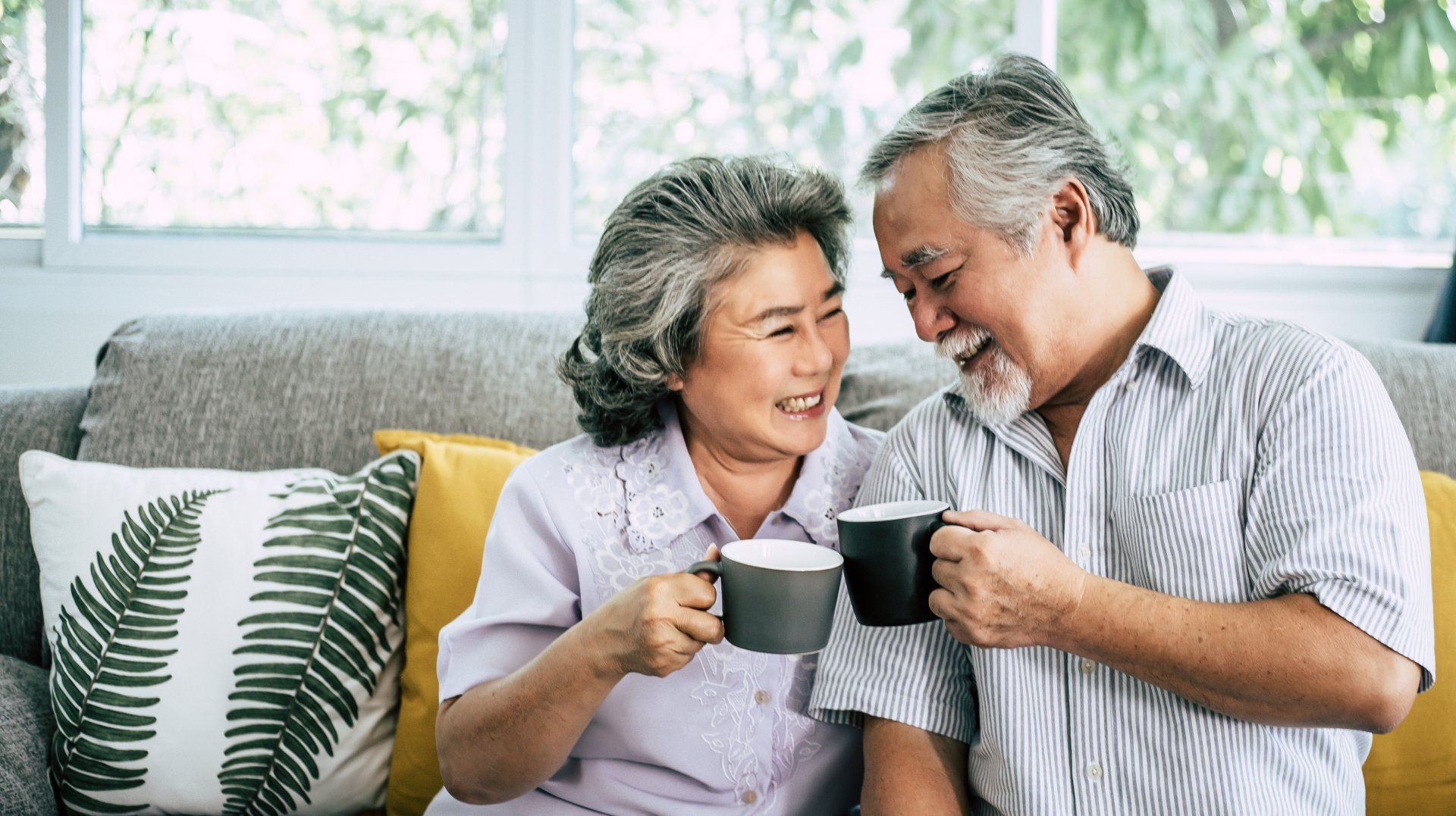 Smiling elderly couple toasting mugs of coffee on a couch.