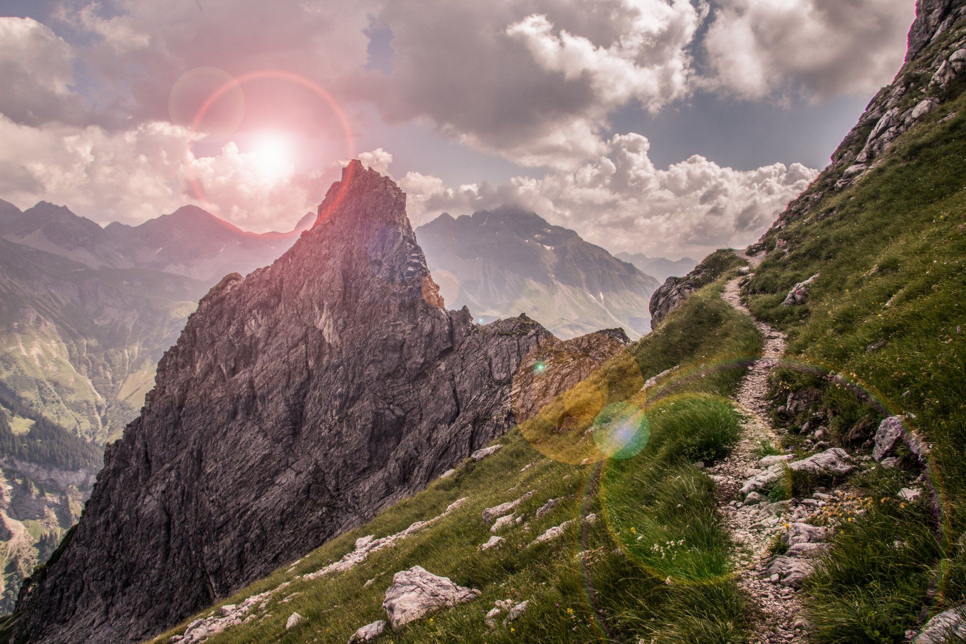 Mountain path winds past a rocky peak towards distant peaks under a cloudy sky with a sun flare.