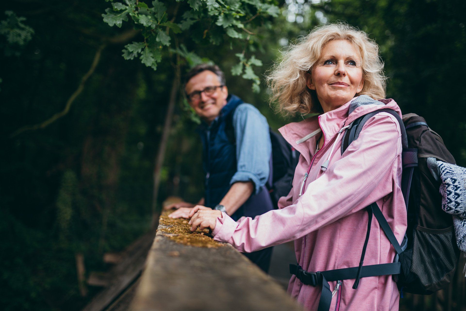 Couple hiking on a wooden bridge in a forest. Woman in pink jacket, man in blue shirt, both smiling.