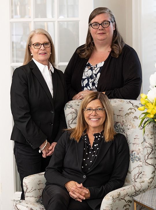 Three professionals in black business attire posed indoors, two standing behind one seated in an armchair.