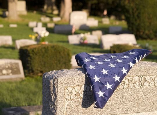 American flag draped over a gravestone in a cemetery with several blurred headstones behind it