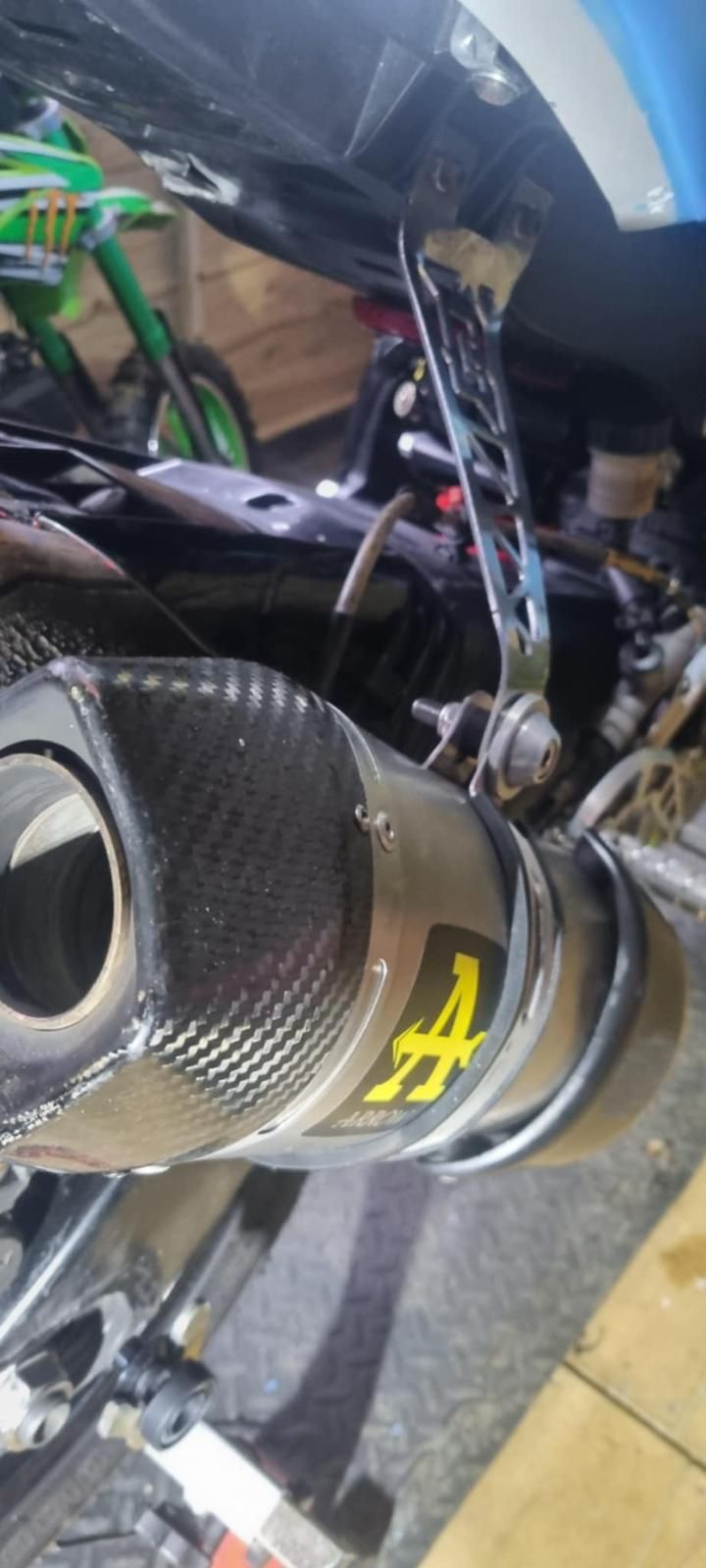 Close-up of a motorcycle exhaust with carbon fiber muffler and yellow logo, under black bodywork.