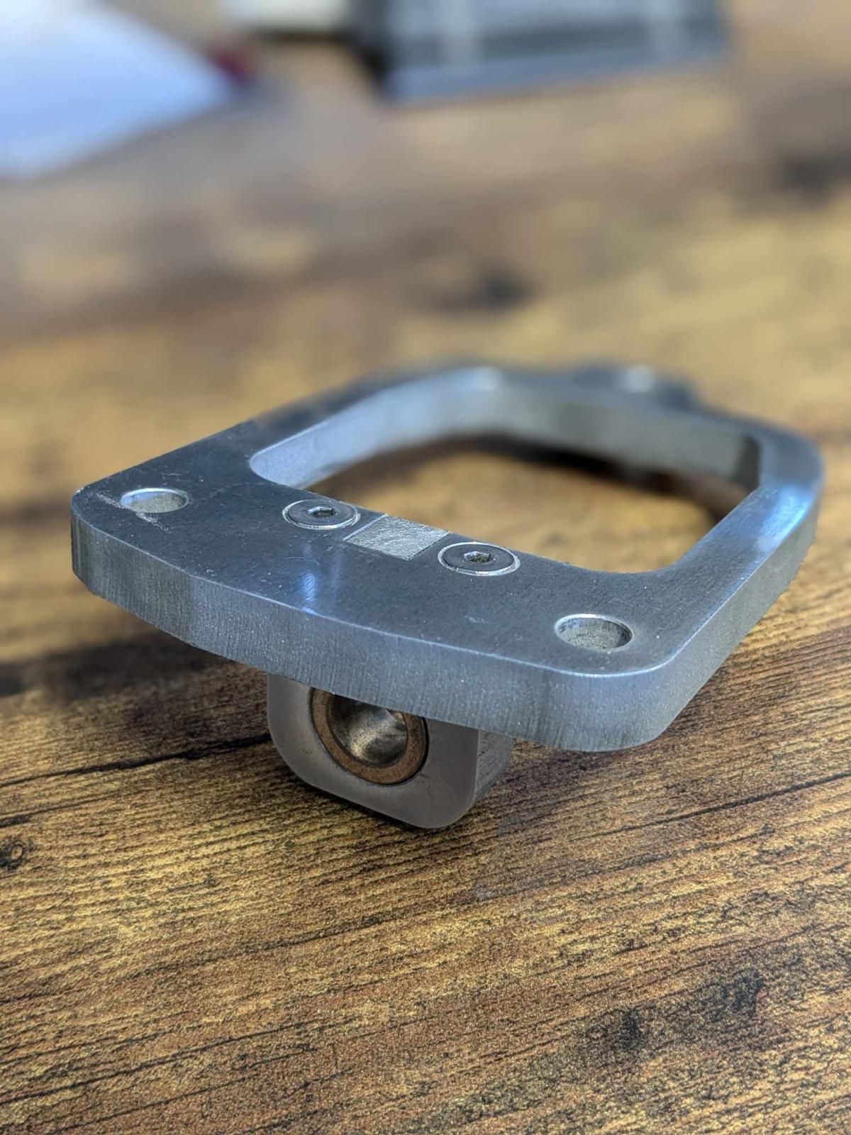Small gray metal bracket with a ring, resting on a wooden surface.