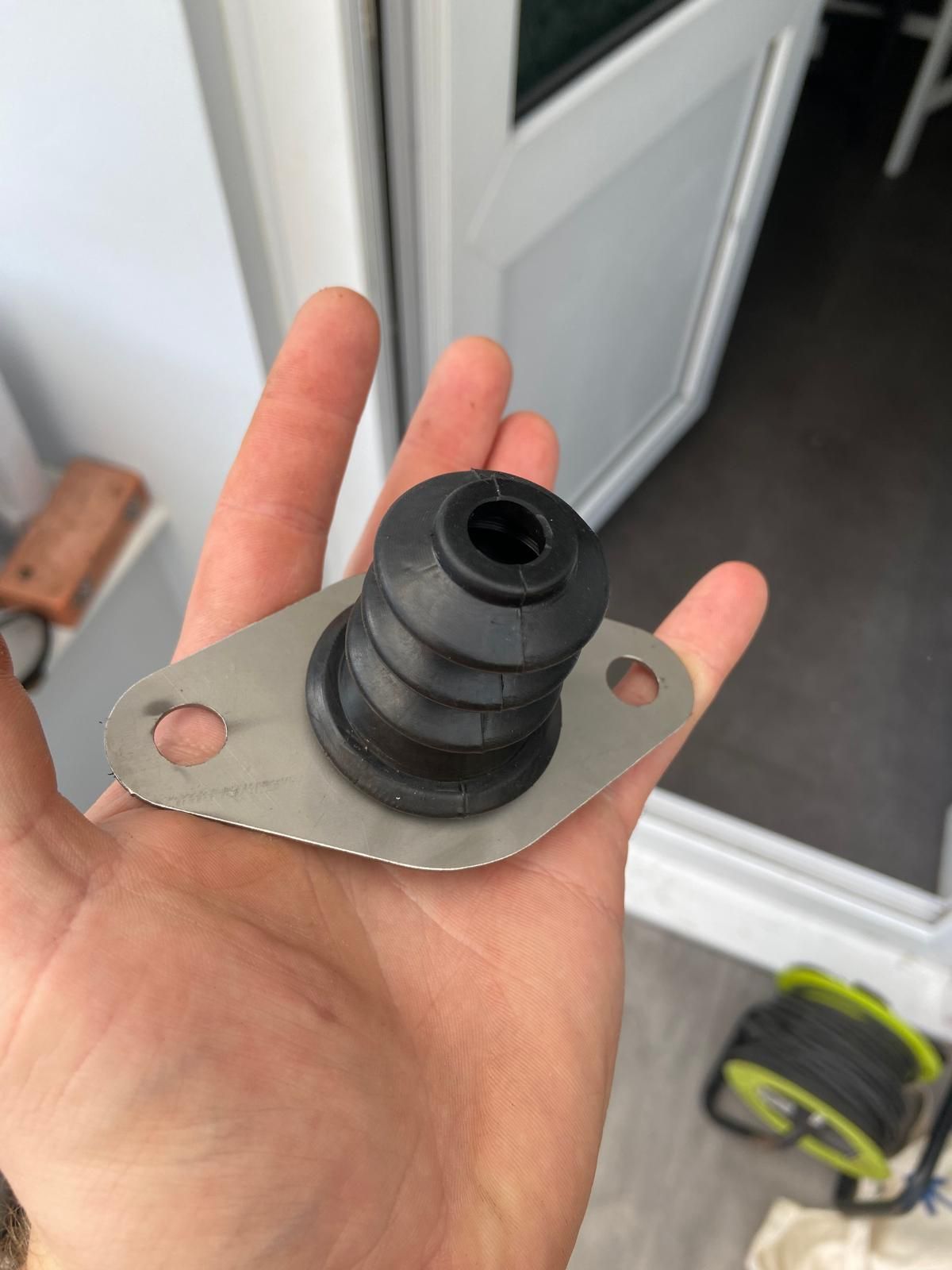Hand holding a black rubber grommet on a metal plate with two bolt holes, indoors near a white door