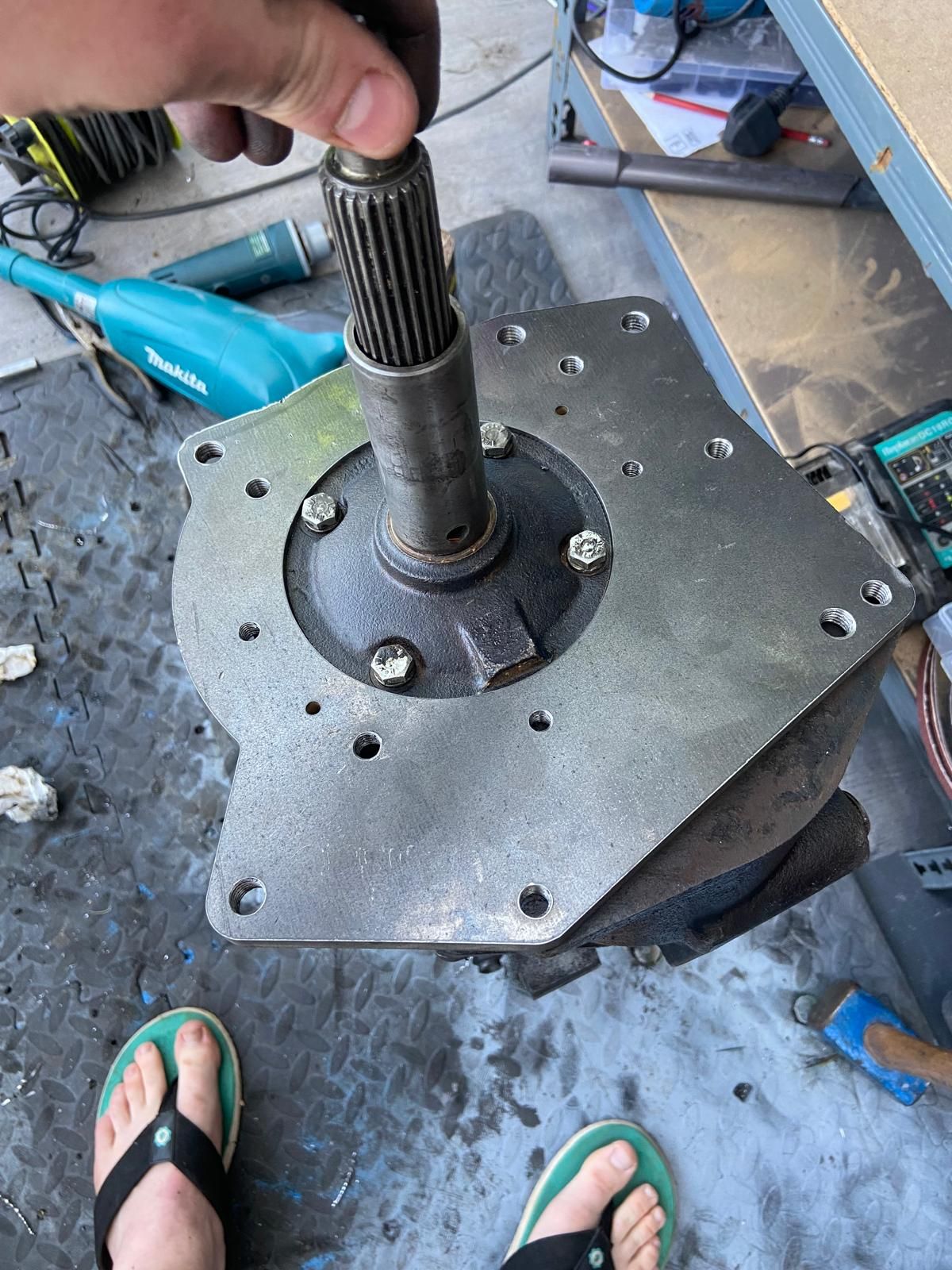 Close-up of a metal machinery shaft and mounting plate on a workshop floor, with a hand holding the splined shaft.