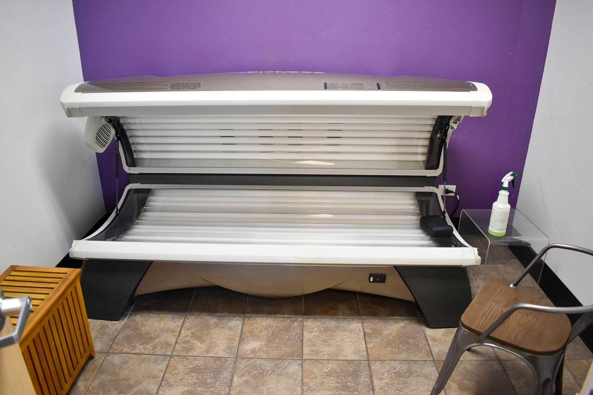 Tanning bed with white shell and black base against a purple wall, with a chair and bottle nearby.