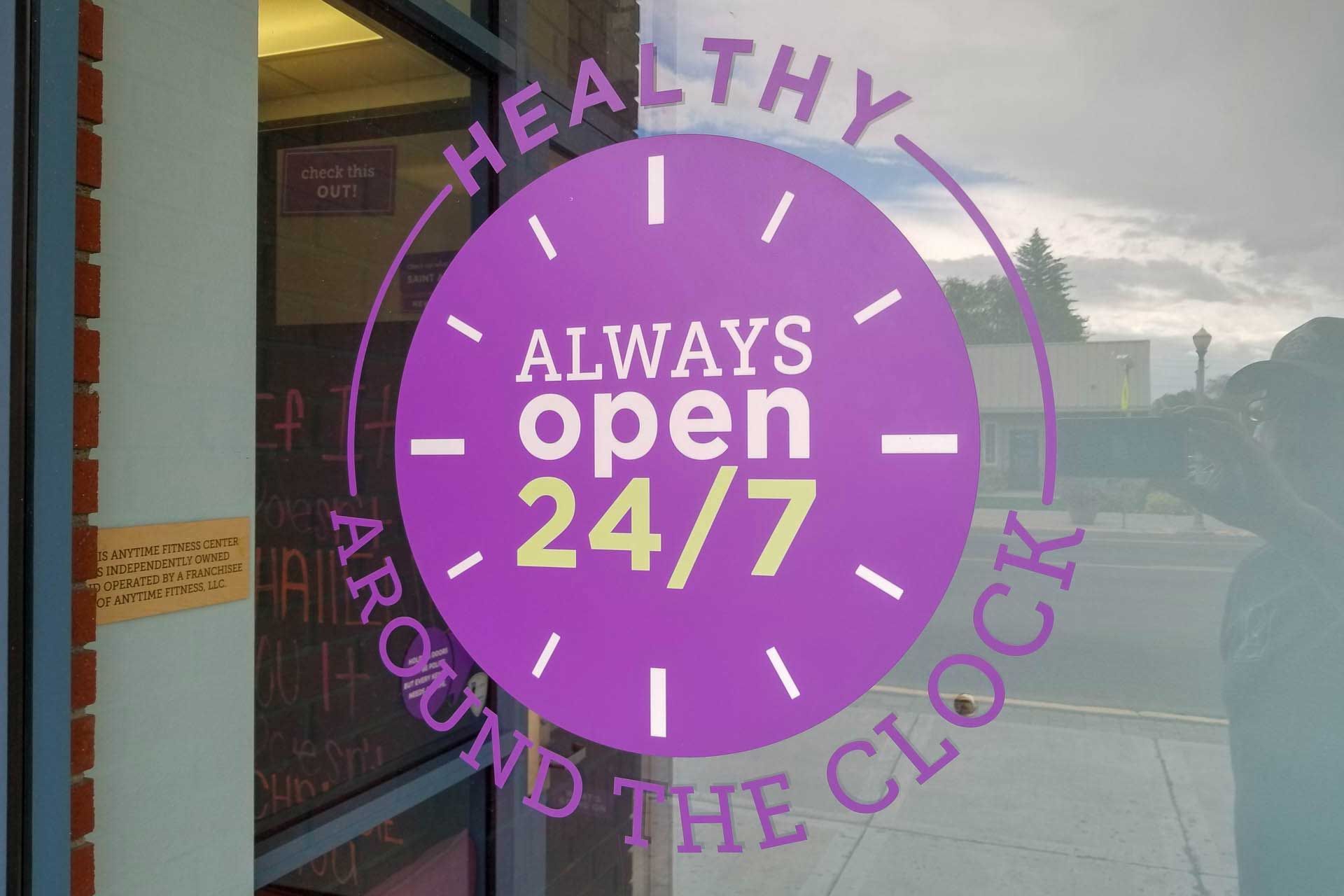 Purple circular “Always open 24/7” sign on a storefront window with “Healthy Around the Clock” text
