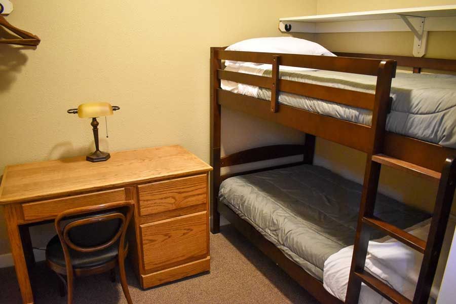 Dorm room with wooden desk, chair, lamp, and a bunk bed with gray bedding.