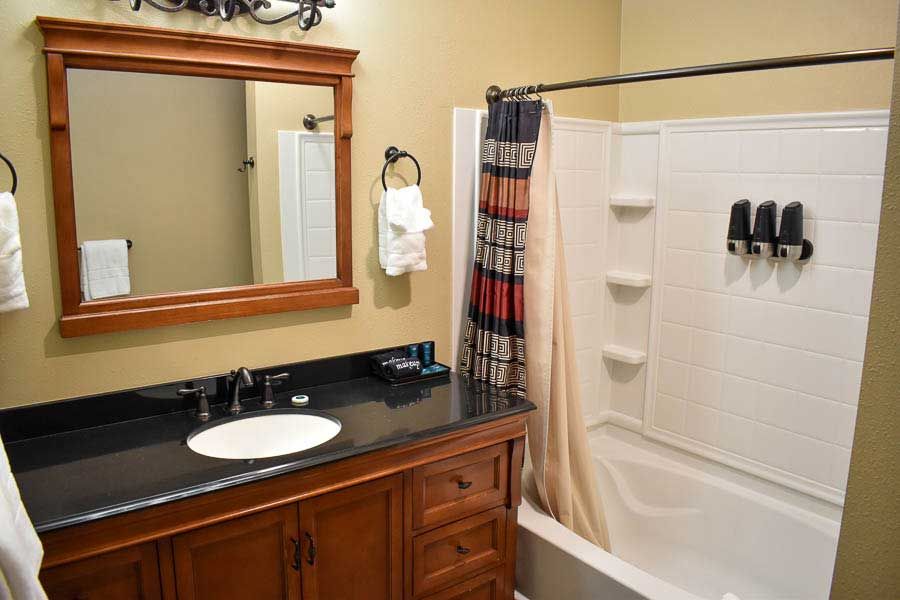 Bathroom with wooden vanity, large mirror, white sink, and white tub with shower curtain and mounted shelves.