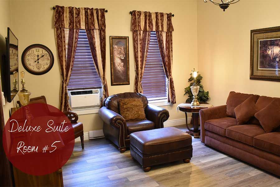 Cozy living room with brown sofas, armchair, ottoman, windows, clock, and red sign reading “Deluxe Suite Room #5”