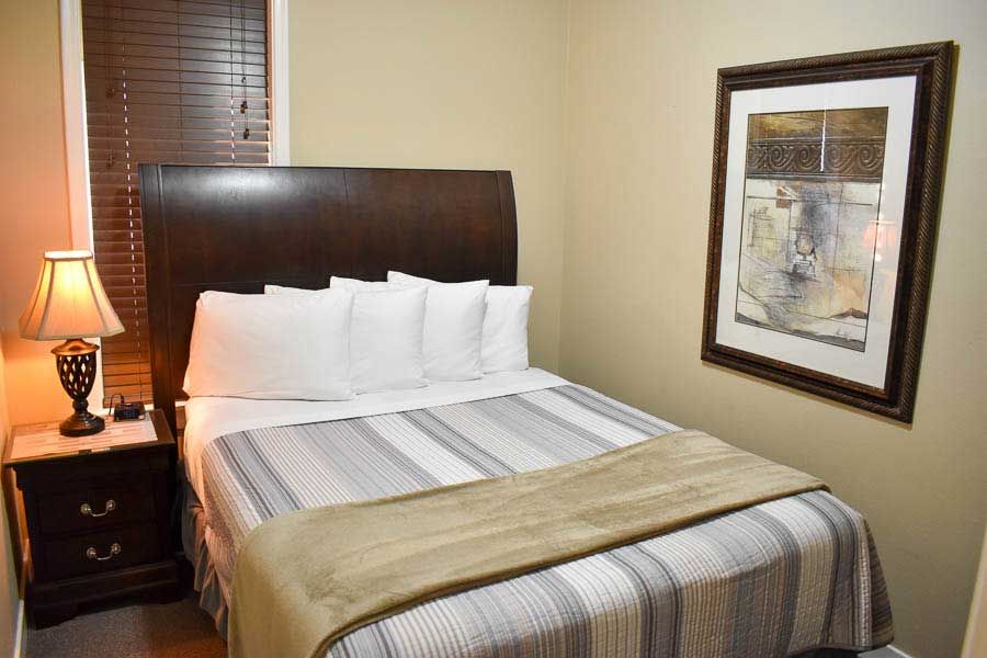 Cozy bedroom with a neatly made bed, dark headboard, lamp, window blinds, and framed wall art.
