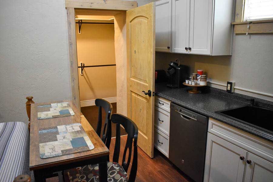 Small kitchen with open wooden door, black countertop, sink, stove, and a narrow table by a bed.