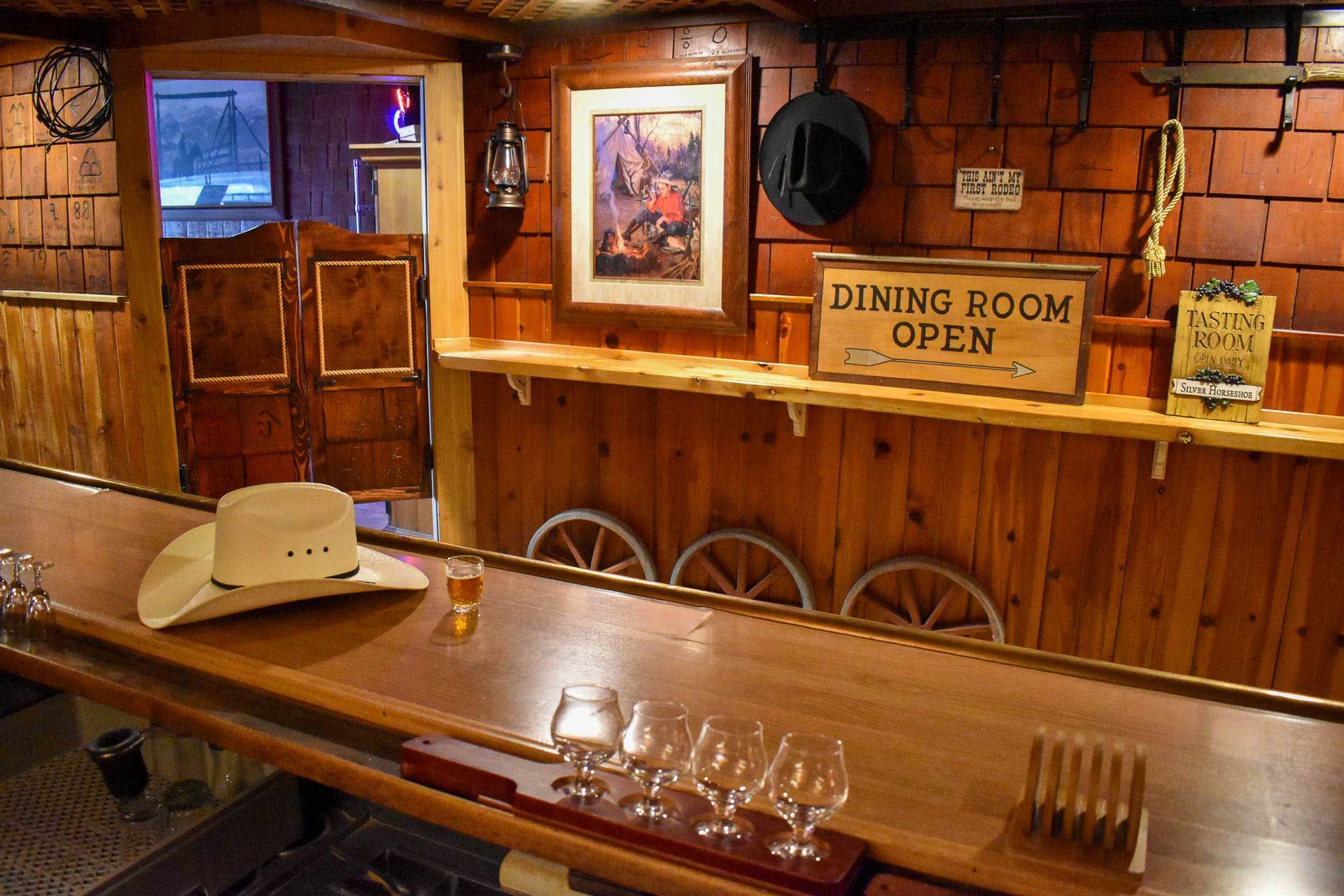 Rustic saloon bar with wooden decor, barstools, a cowboy hat, and a sign reading “Dining Room Open.”
