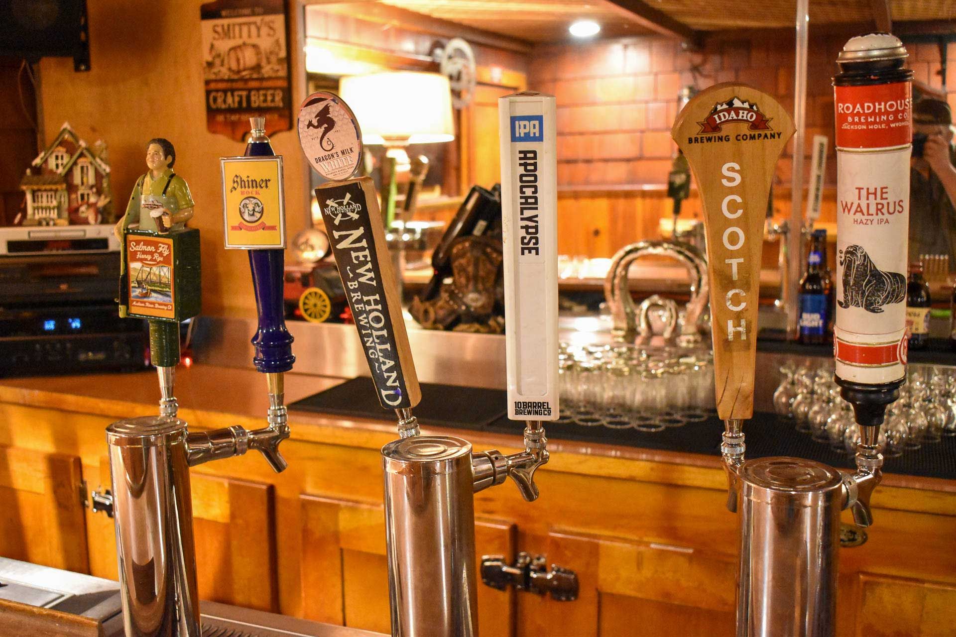 Beer taps lined up on a wooden bar in a warmly lit pub, with branded tap handles and bottles behind them
