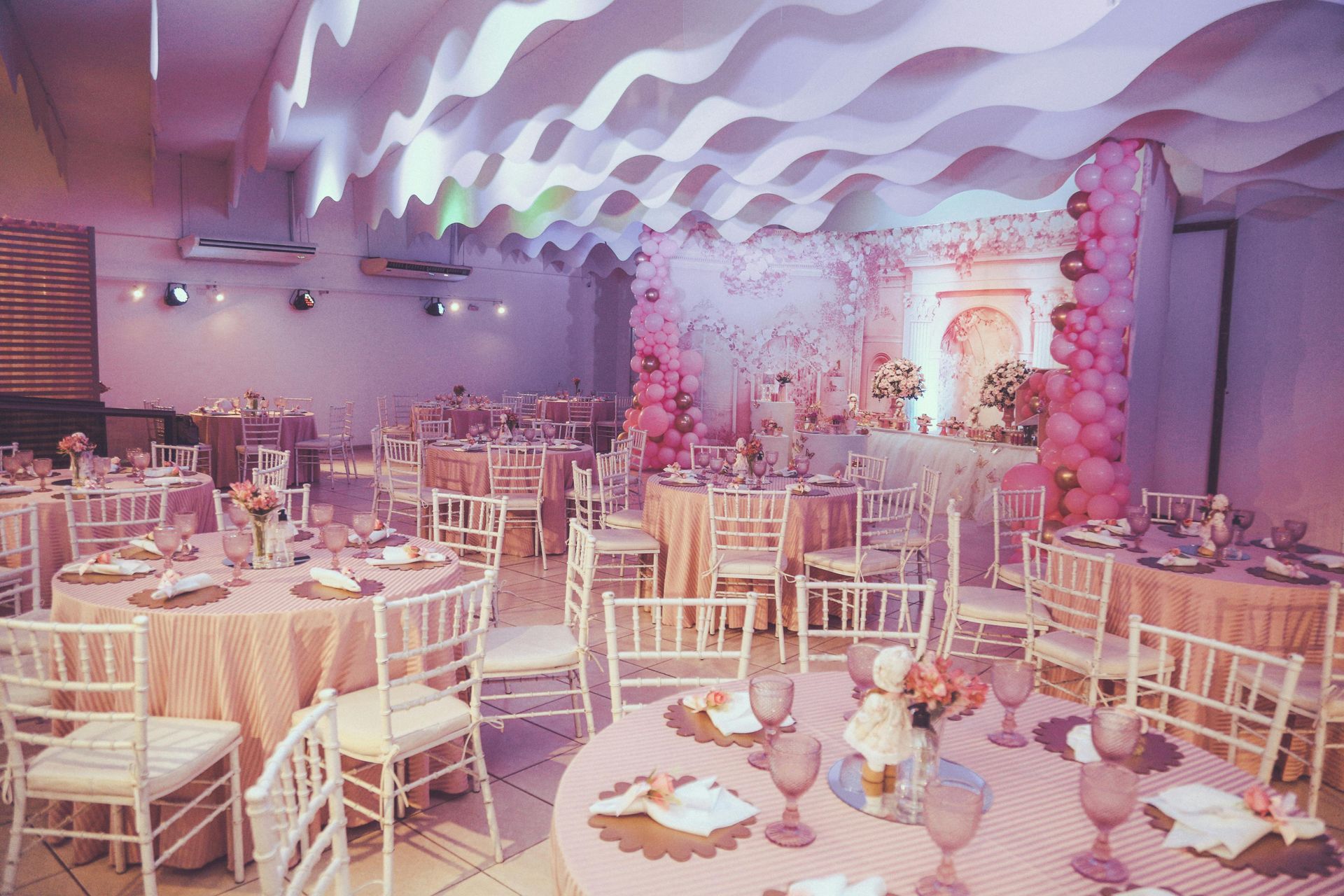 Decorated banquet hall with round tables, white chairs, pink floral decor, and wavy ceiling lights