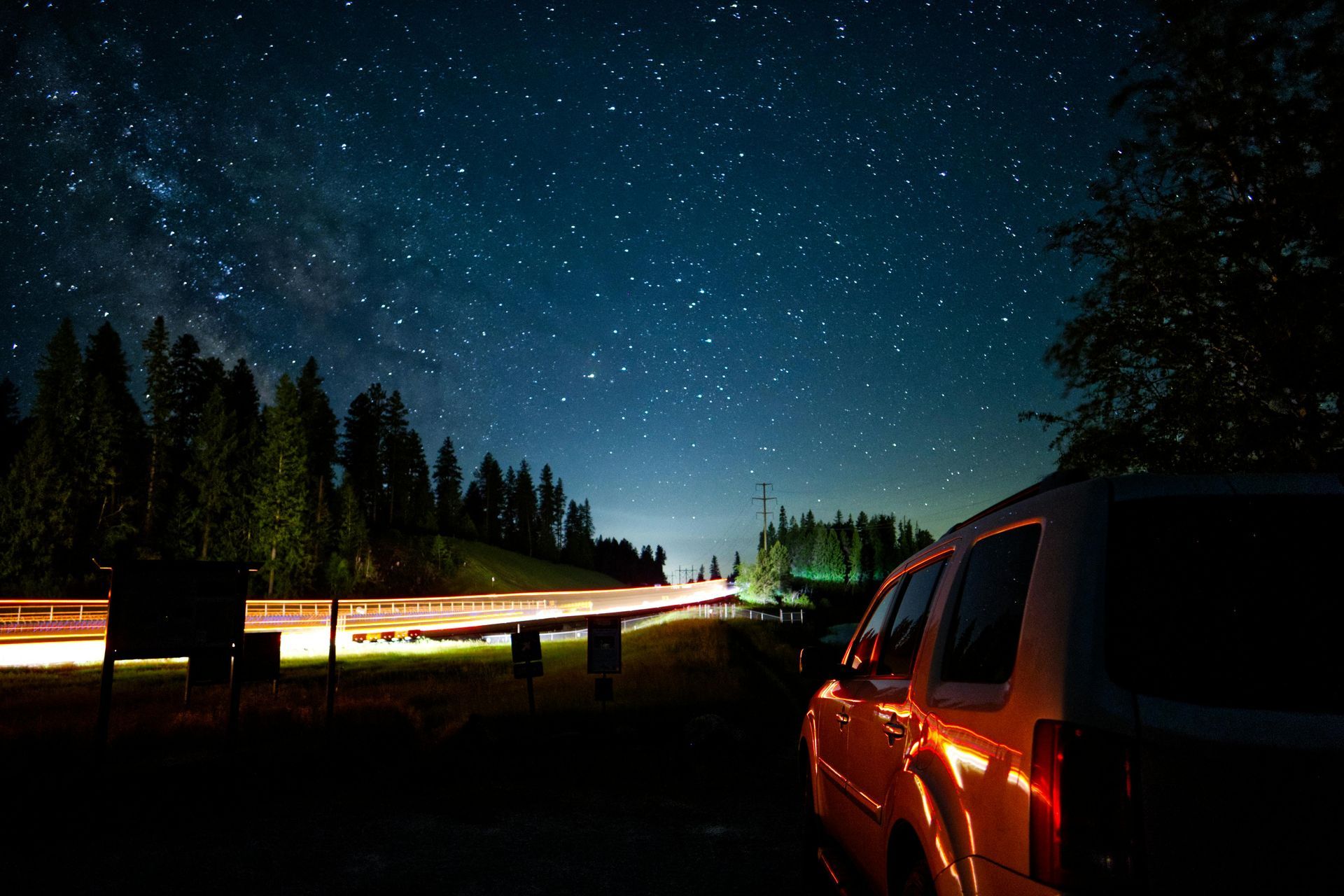 Night sky over a forest road with a car at right and light trails from passing traffic