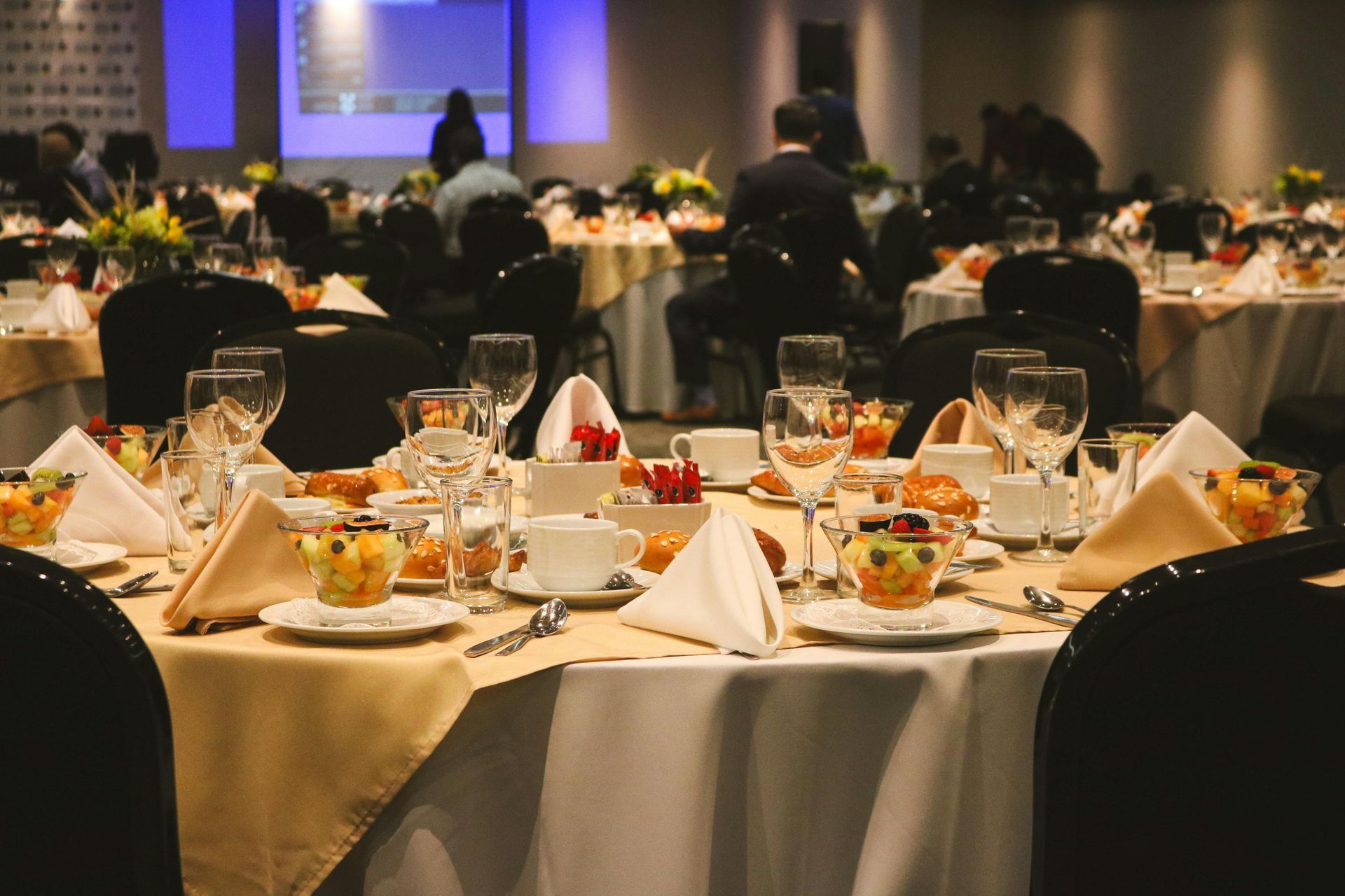 Banquet tables set for a formal event in a dimly lit conference hall, with a stage in the background