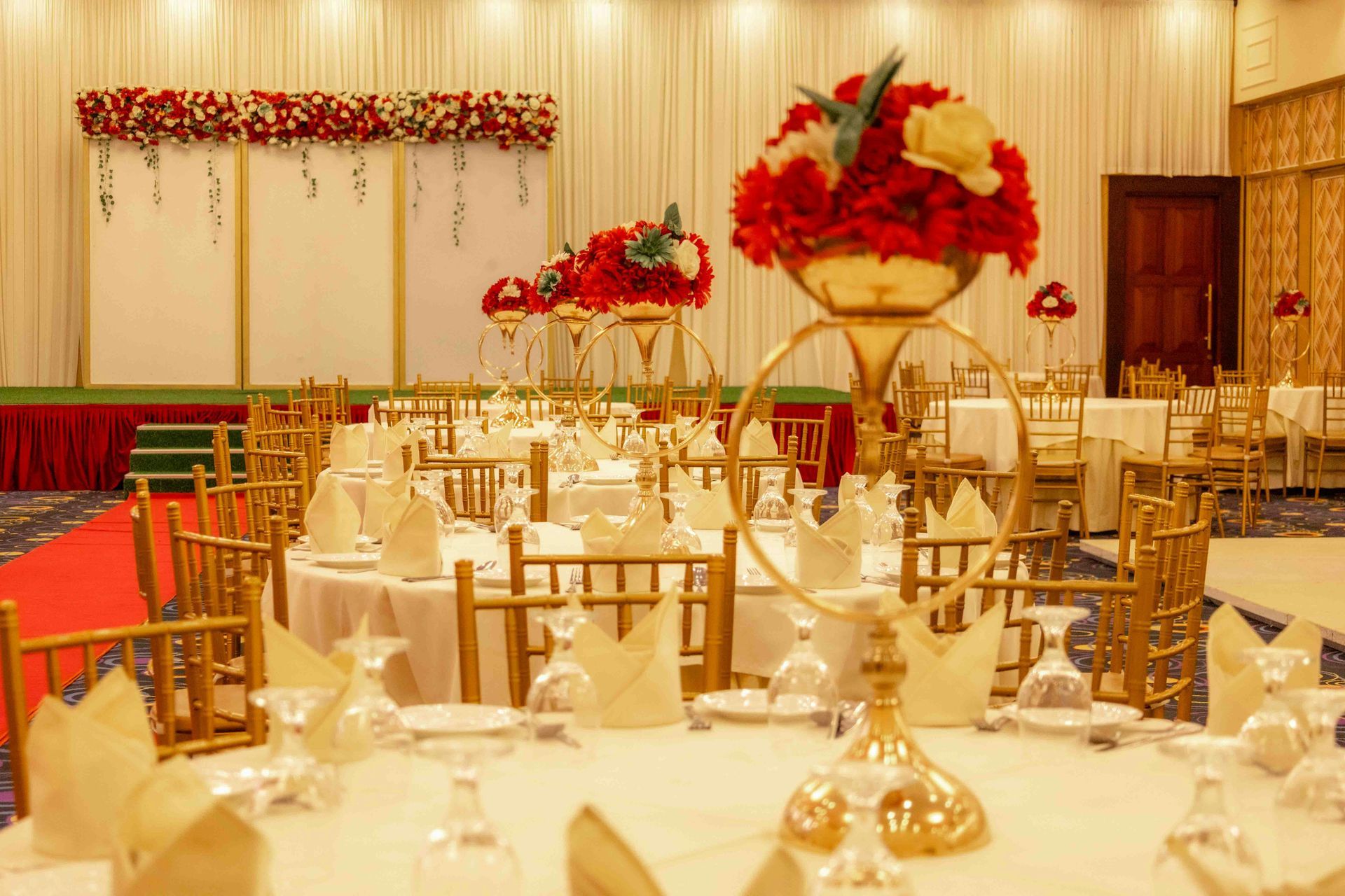 Elegant banquet hall with white table settings, gold chairs, and red floral centerpieces under warm lighting