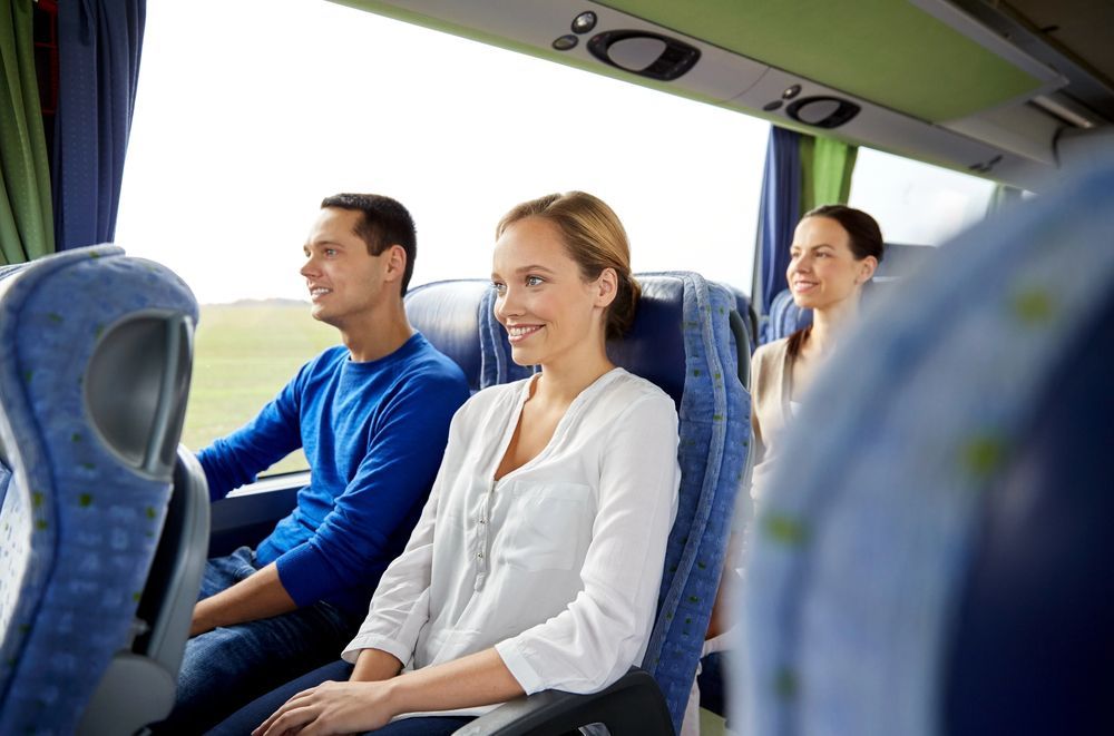 Tres personas sentadas en un autobús, sonriendo, mirando por las ventanas. Asientos verdes y azules. Luz natural brillante.
