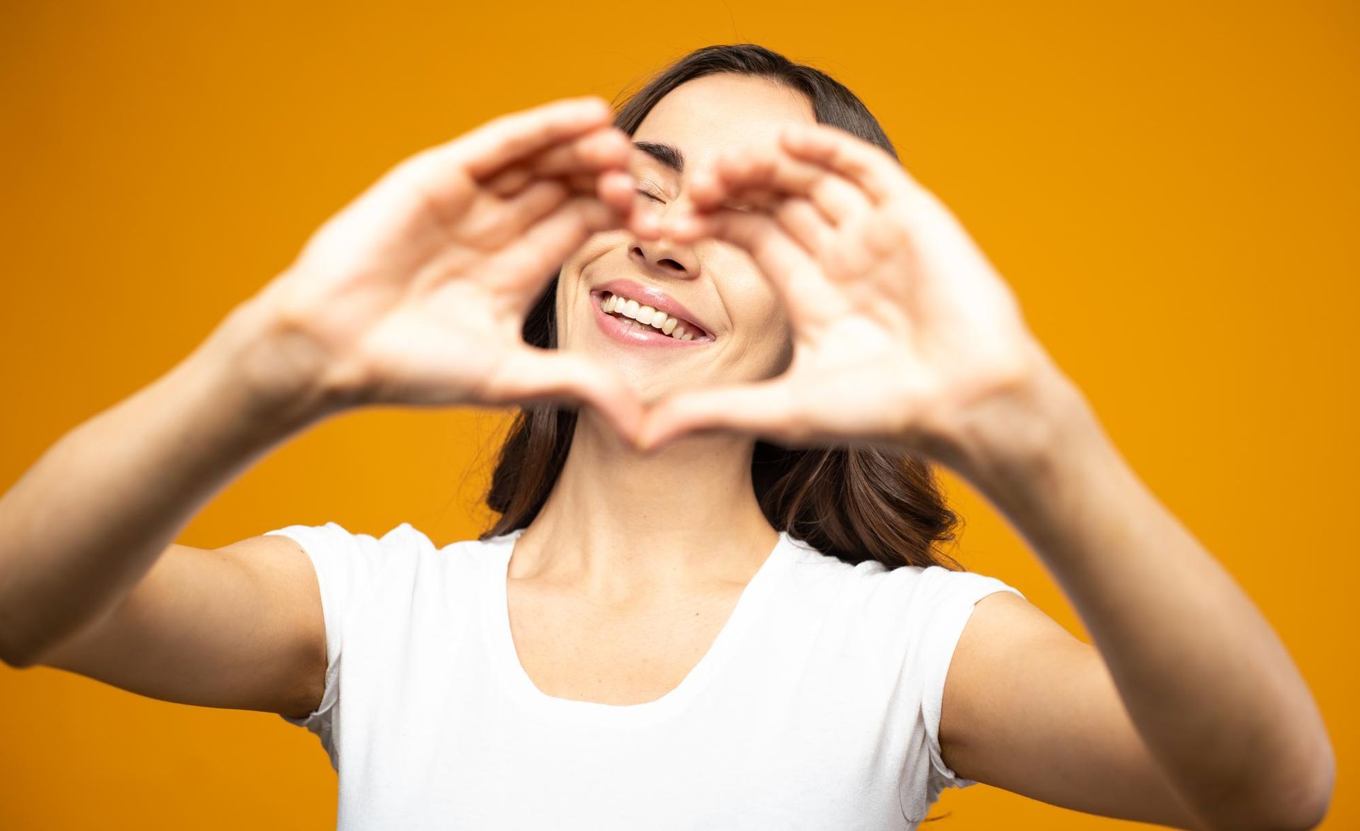 Smiling person in a white shirt making a heart shape with hands against an orange background