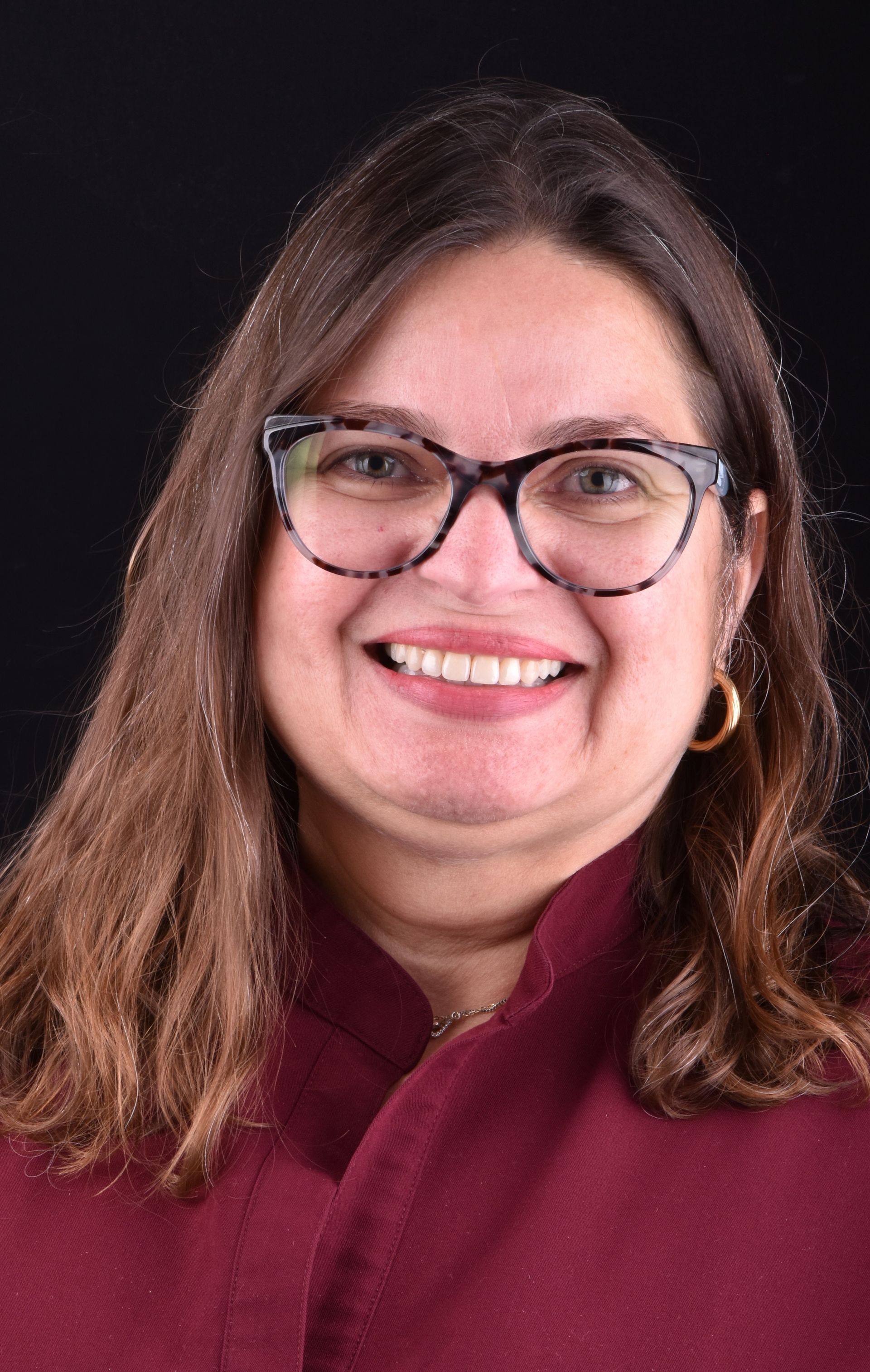 Smiling person with glasses and long brown hair wearing a maroon sweater against a dark background