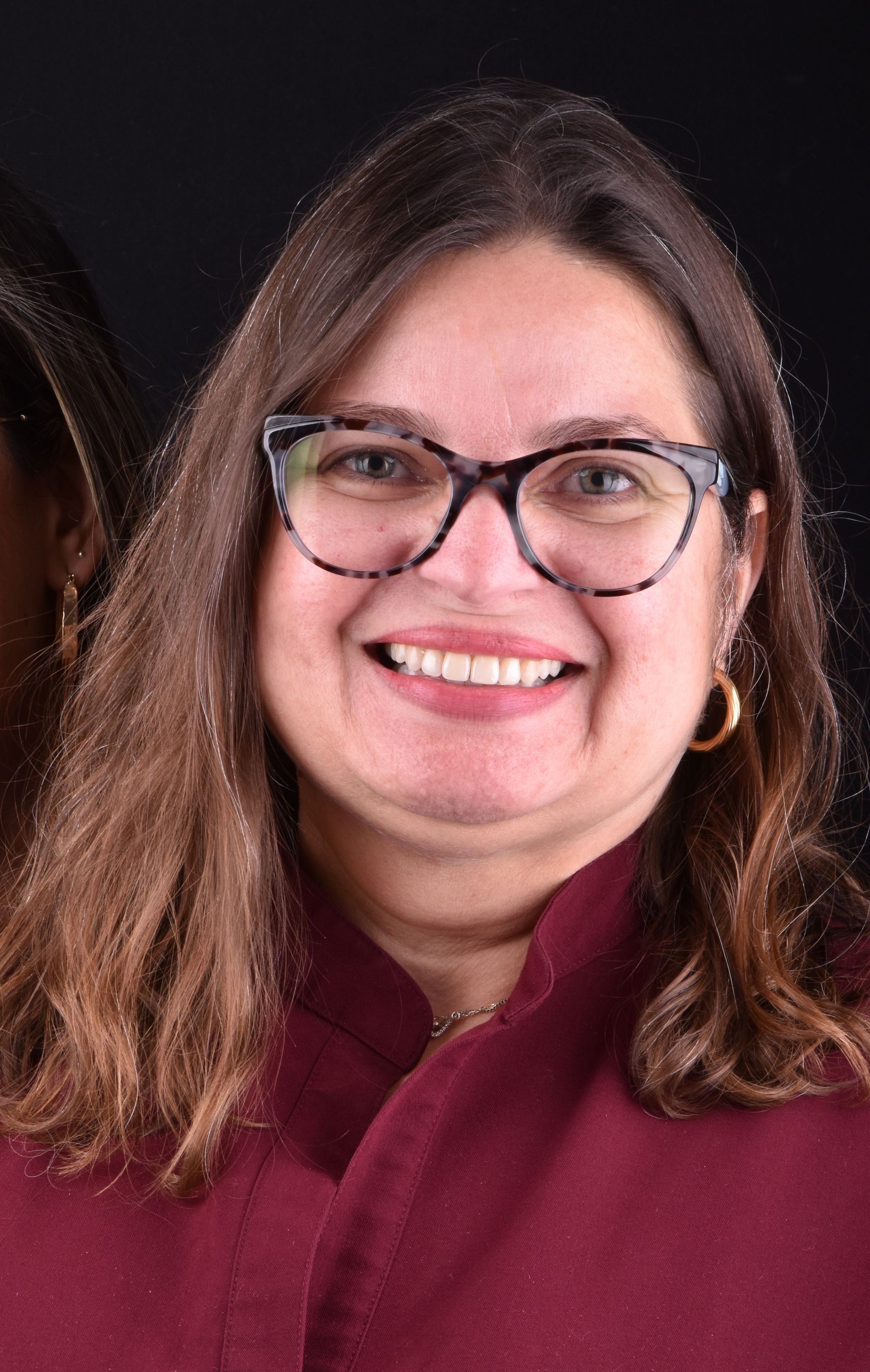 Smiling person with glasses and long brown hair wearing a maroon sweater against a dark background