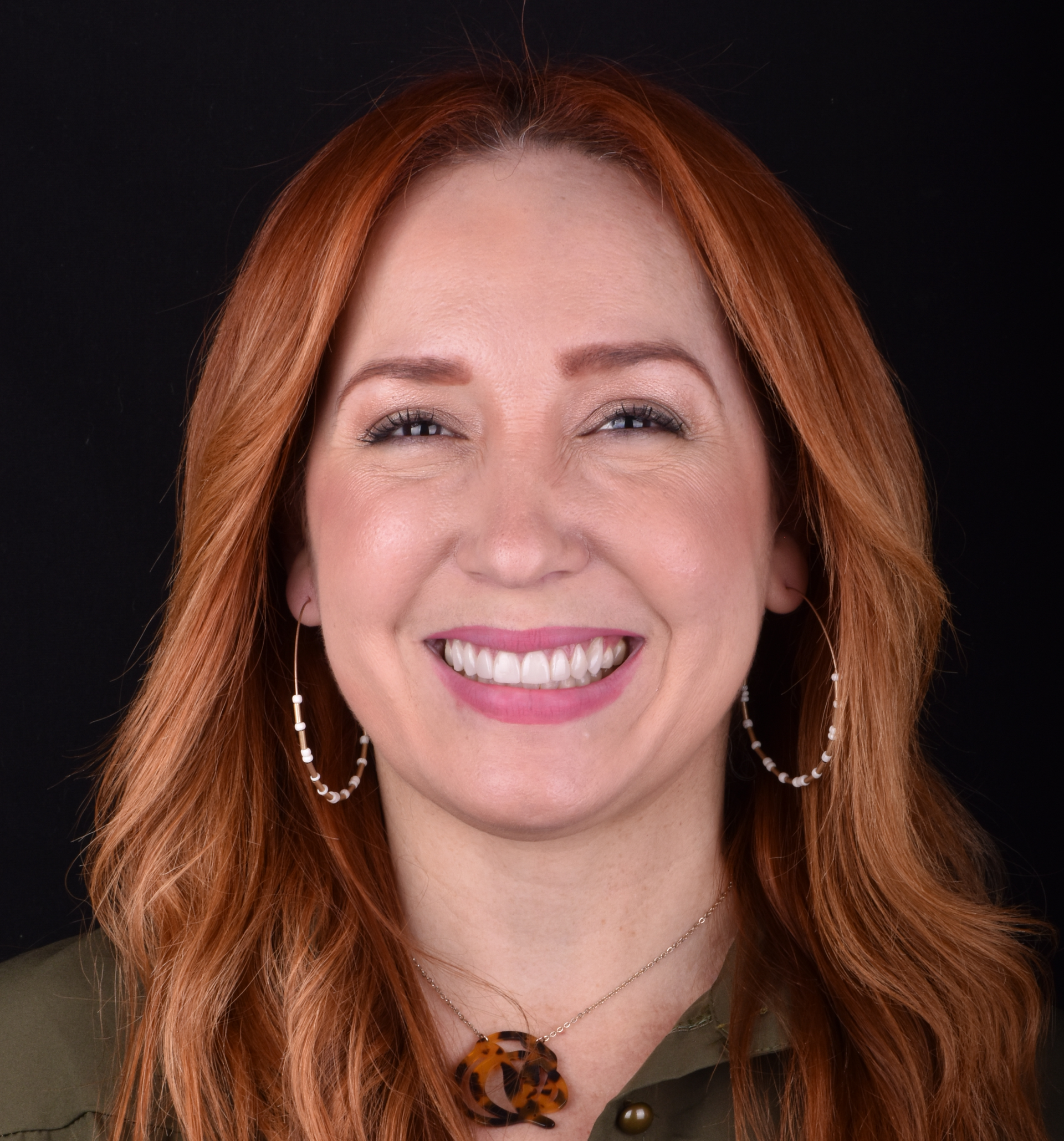Smiling woman with red hair, hoop earrings, and an olive top against a black background