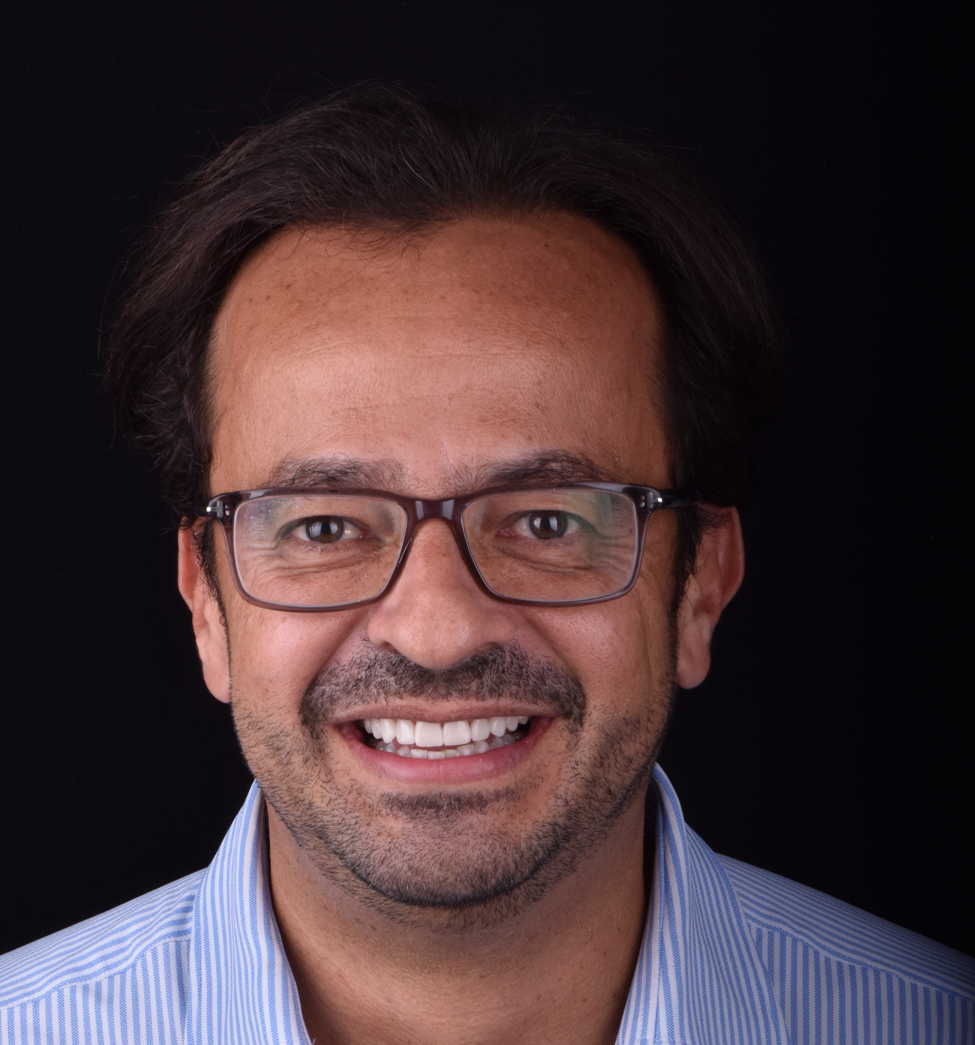 Smiling man with glasses against a dark background, wearing a light blue shirt