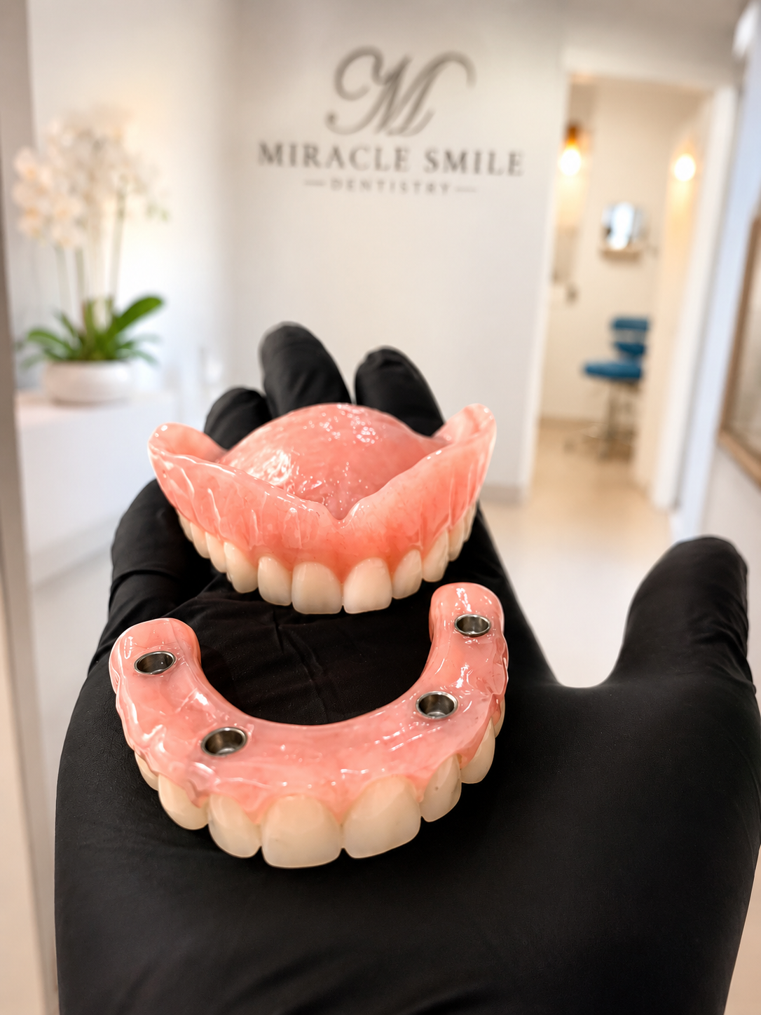 Gloved hand holding two pink dentures in a dental clinic with a “Miracle Smile” sign in the background