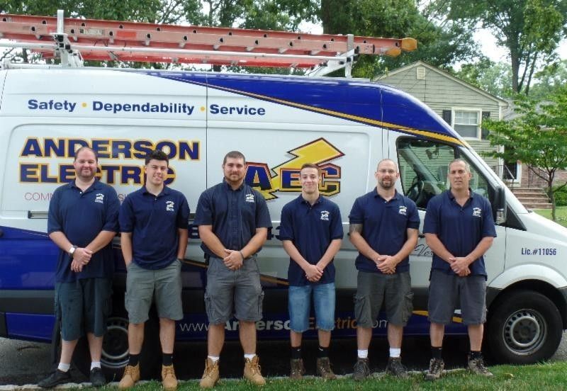Six employees in navy uniforms stand in front of an Anderson Electric company van parked outdoors.