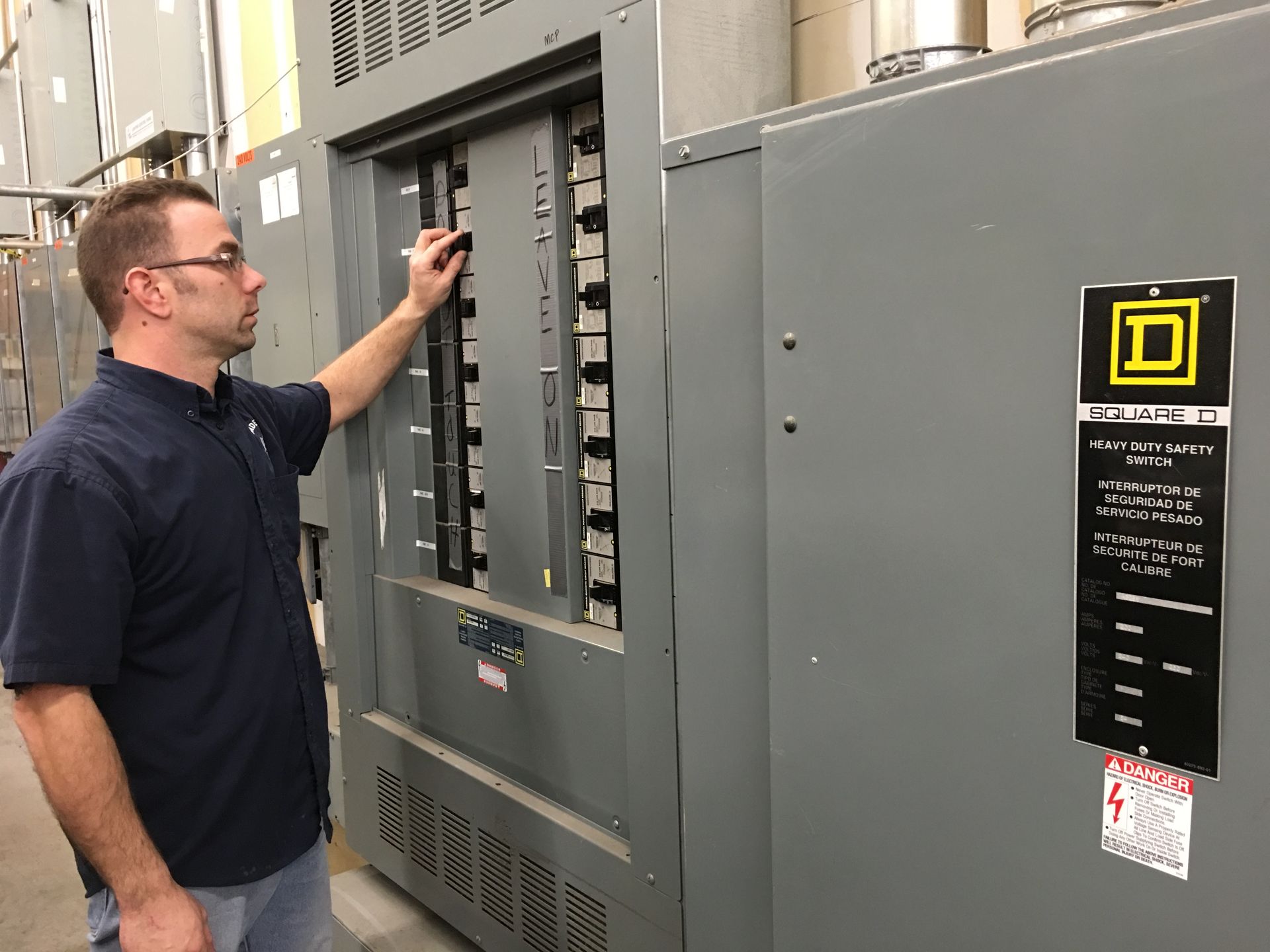 A person in a blue shirt adjusting a switch on a large grey industrial electrical panel marked with a Square D logo.