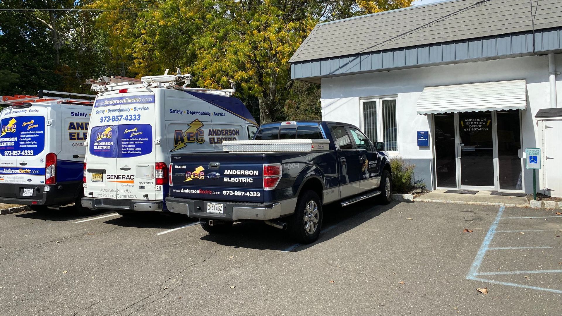 Two white and blue American Electric work vans and a dark blue pickup truck parked in front of a white commercial building.