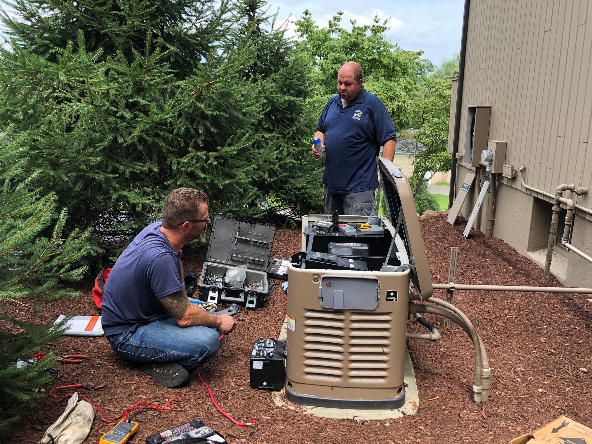 Two technicians work on an open outdoor standby generator next to a house wall and evergreen trees.