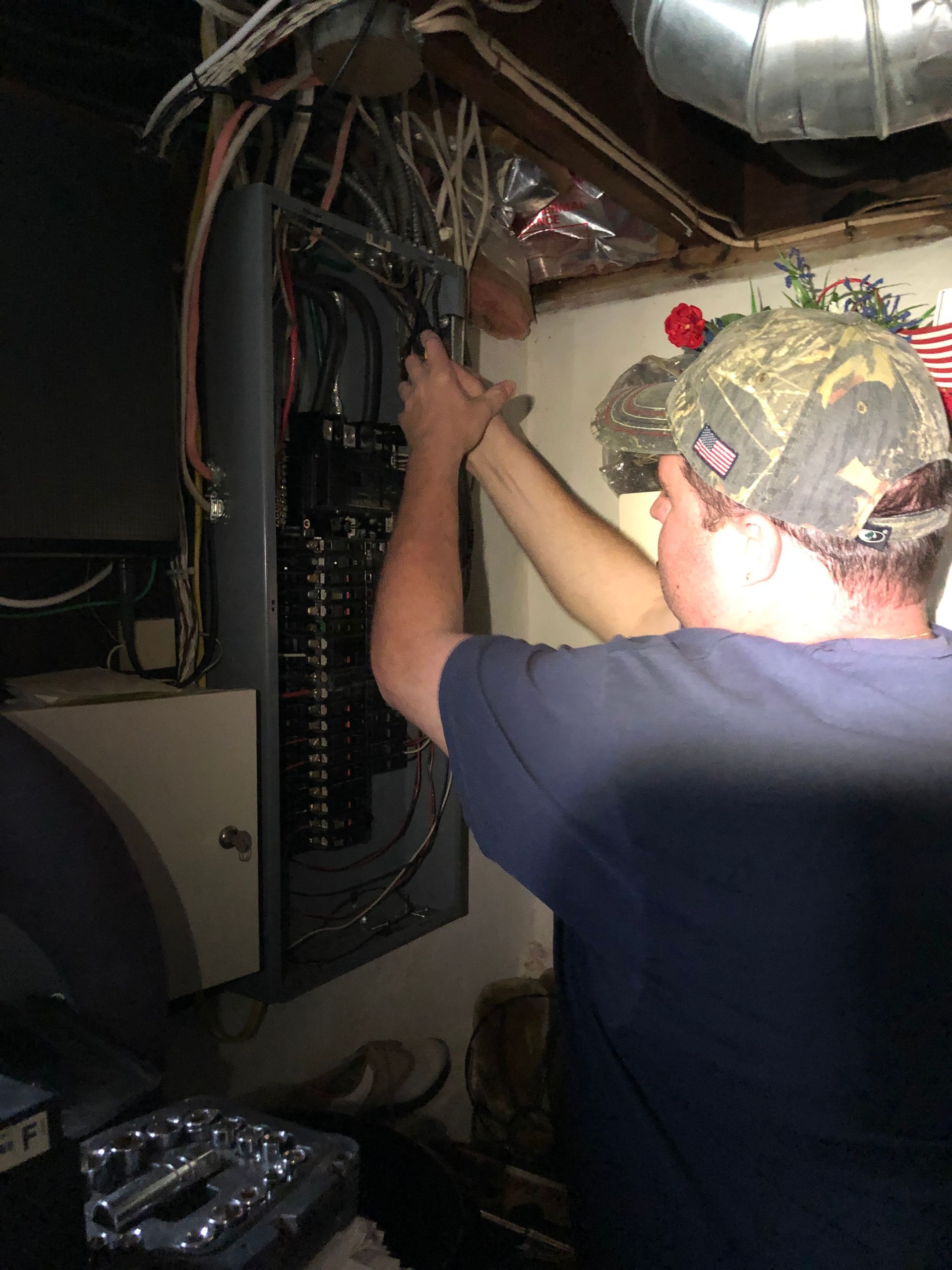 A person in a camouflage cap works on the wiring inside an open electrical breaker panel in a dimly lit space.