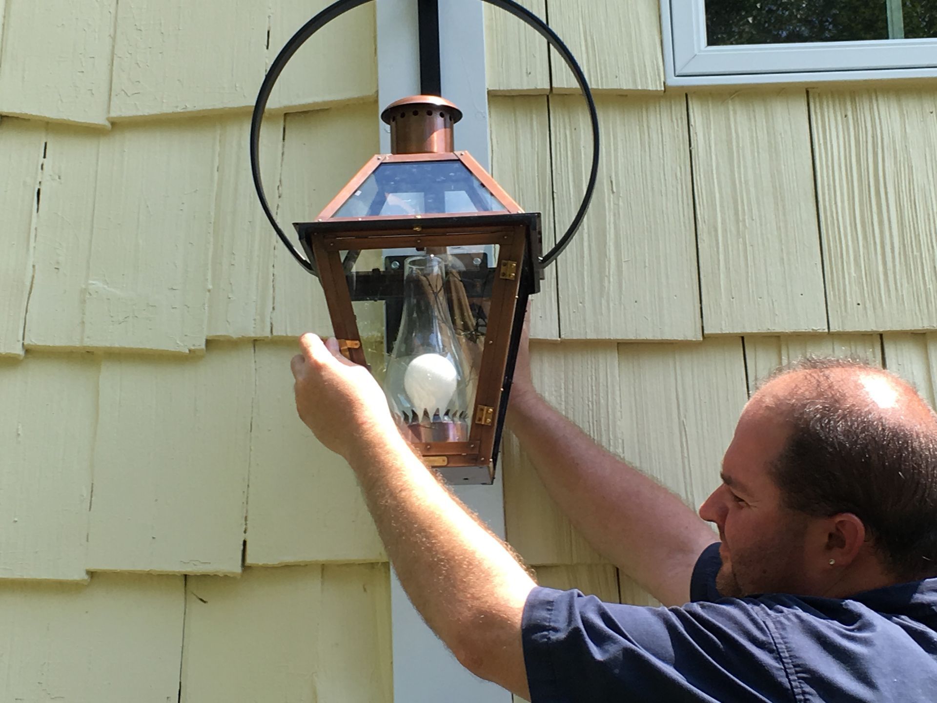 A person installing or adjusting a copper and glass outdoor wall lantern on a yellow shingled exterior wall.