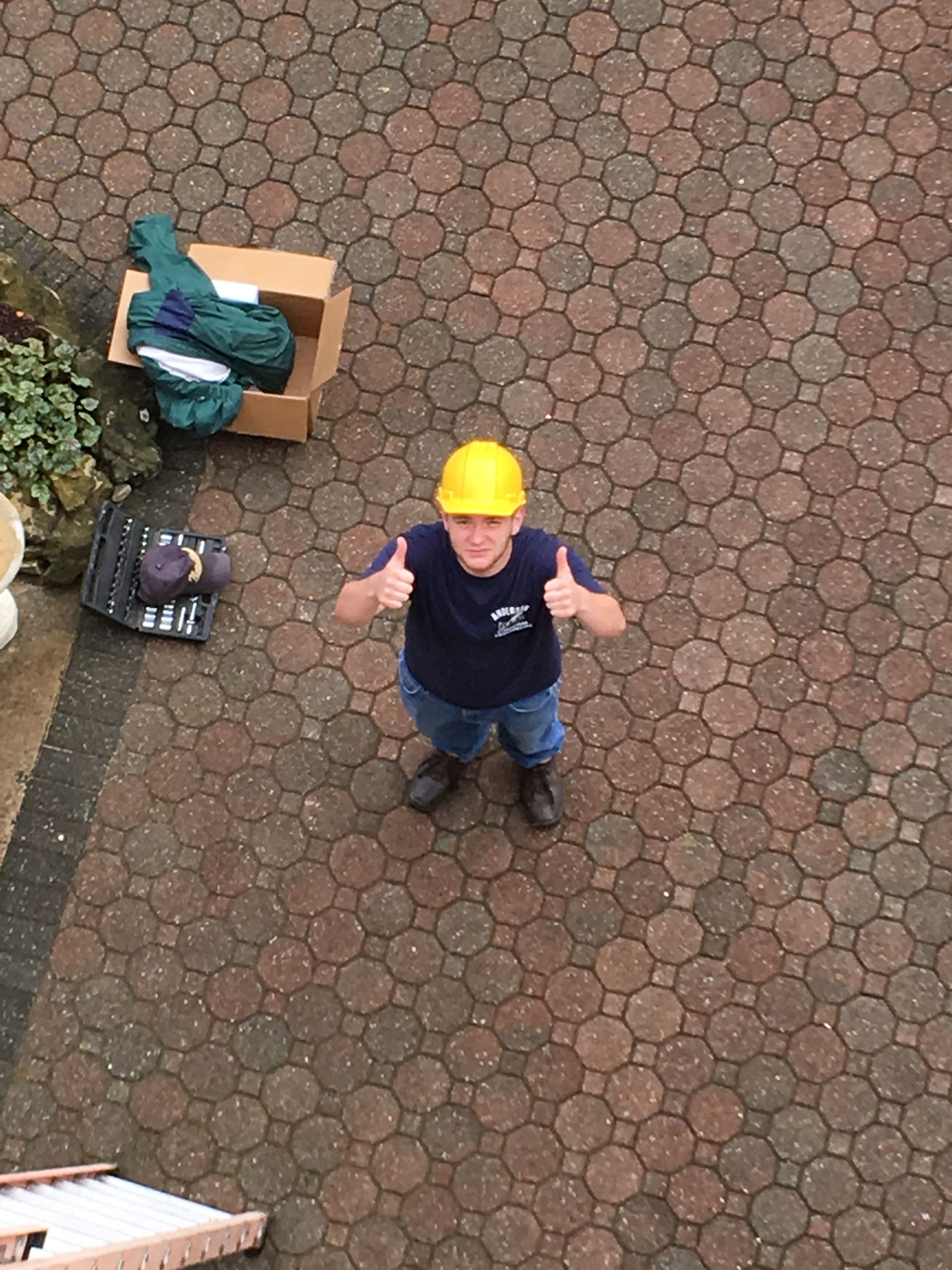 A person wearing a yellow hard hat stands on a brick patio, giving a thumbs up with both hands.