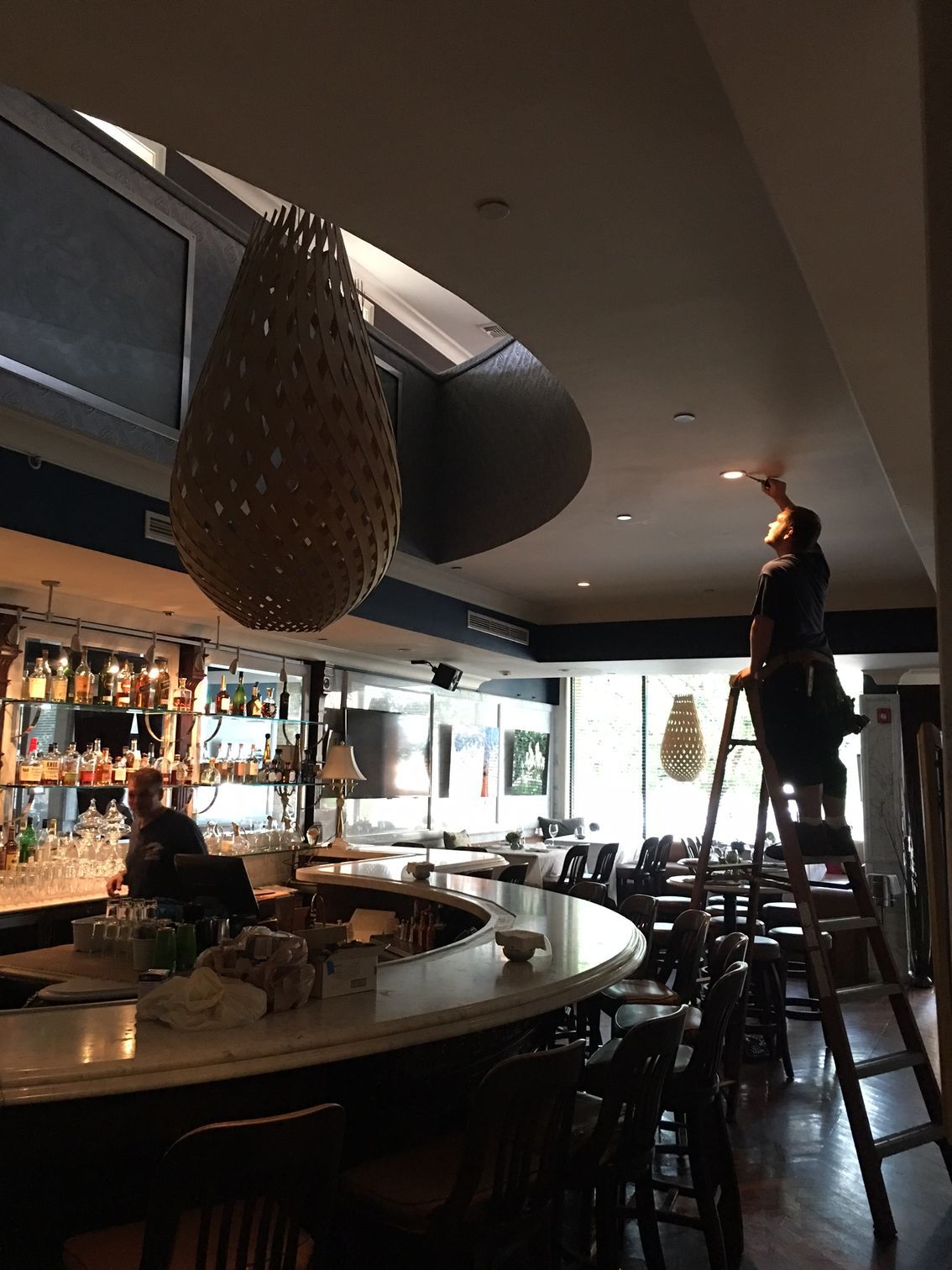 A person stands on a tall ladder, repairing a ceiling light in a dimly lit restaurant with a curved bar.